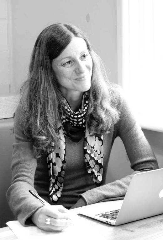 Black and white photo of a woman with long hair, sitting at a table, looking out the window, holding a pen, with an open laptop in front of her, in an indoor setting.