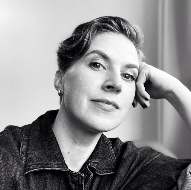 Black and white portrait of a woman with short hair, resting her head on her hand, wearing a denim jacket, looking at the camera with a relaxed expression.