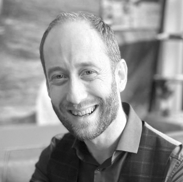 Black and white photo of a smiling man with short hair and a beard, wearing a collared shirt and vest, sitting indoors with a wooden wall in the background.