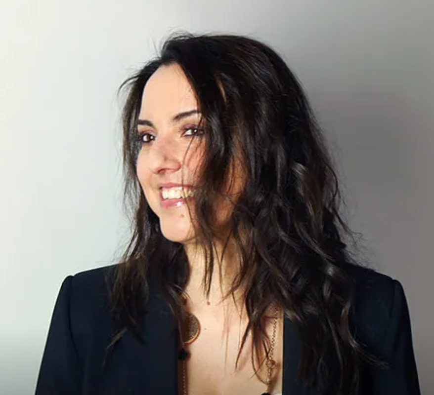 Woman with shoulder-length curly dark hair, smiling and looking to the side, wearing a black blazer and a gold necklace.