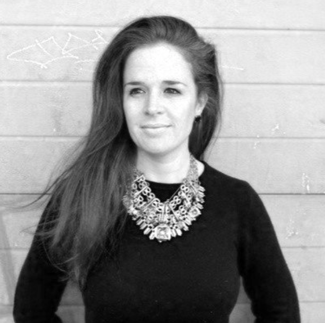Black and white photo of a woman with long hair, wearing a statement necklace and a black sweater, standing outdoors in front of a wooden wall.