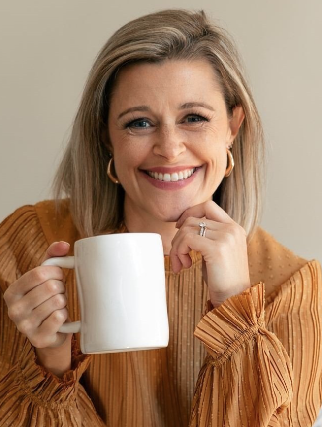 A woman smiling while holding a white mug, wearing an orange striped blouse with ruffled sleeves, and earrings.