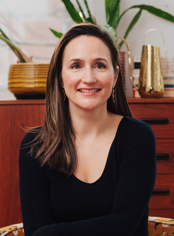 A woman with long brown hair, wearing a black long-sleeve shirt and gold earrings, sitting in a room with wooden furniture, plants, and decorative items in the background.
