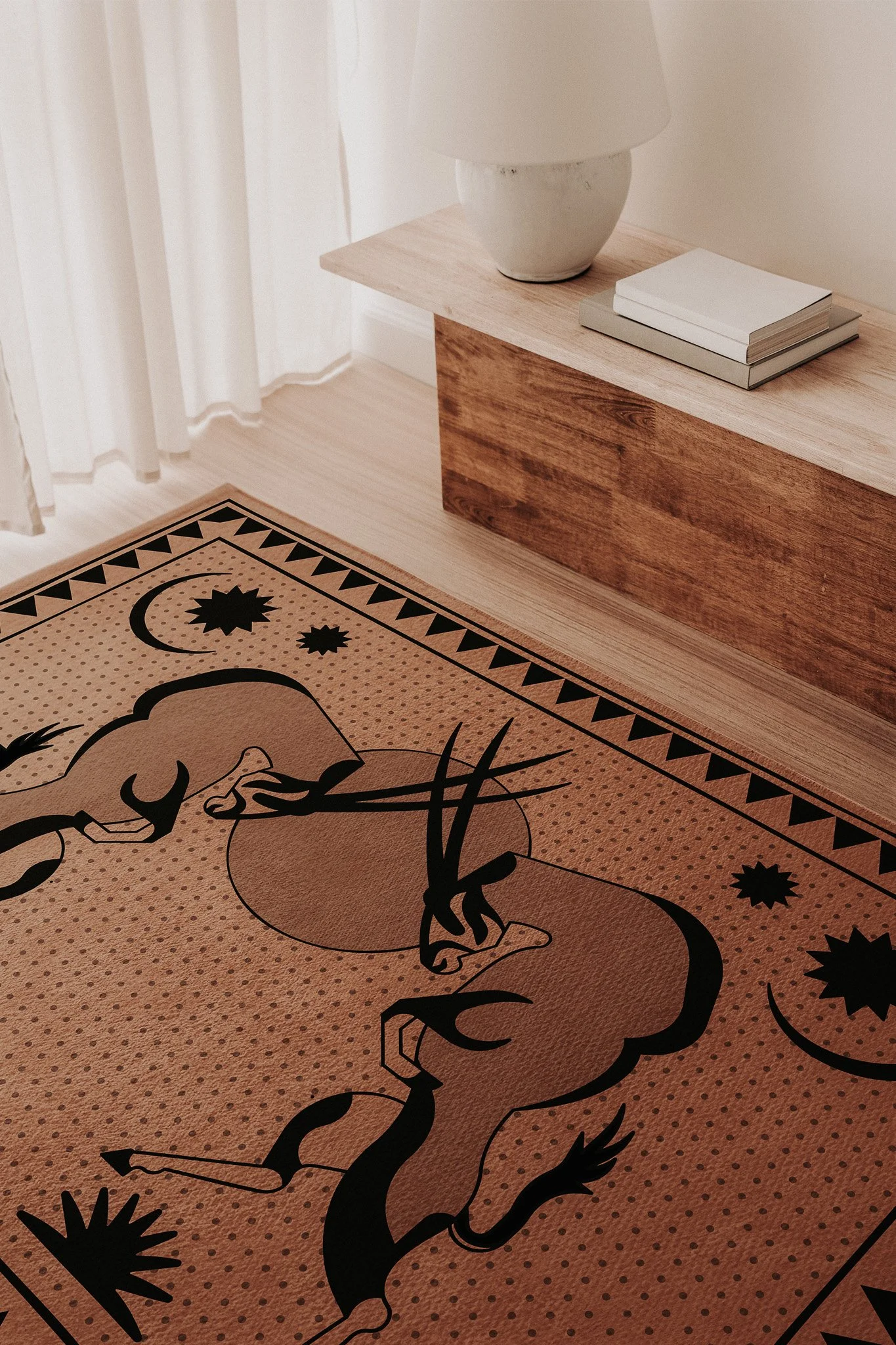 Close-up of a beige area rug with black tribal-style oryx and celestial designs, placed next to a wooden side table with a white ceramic lamp and stacked books.