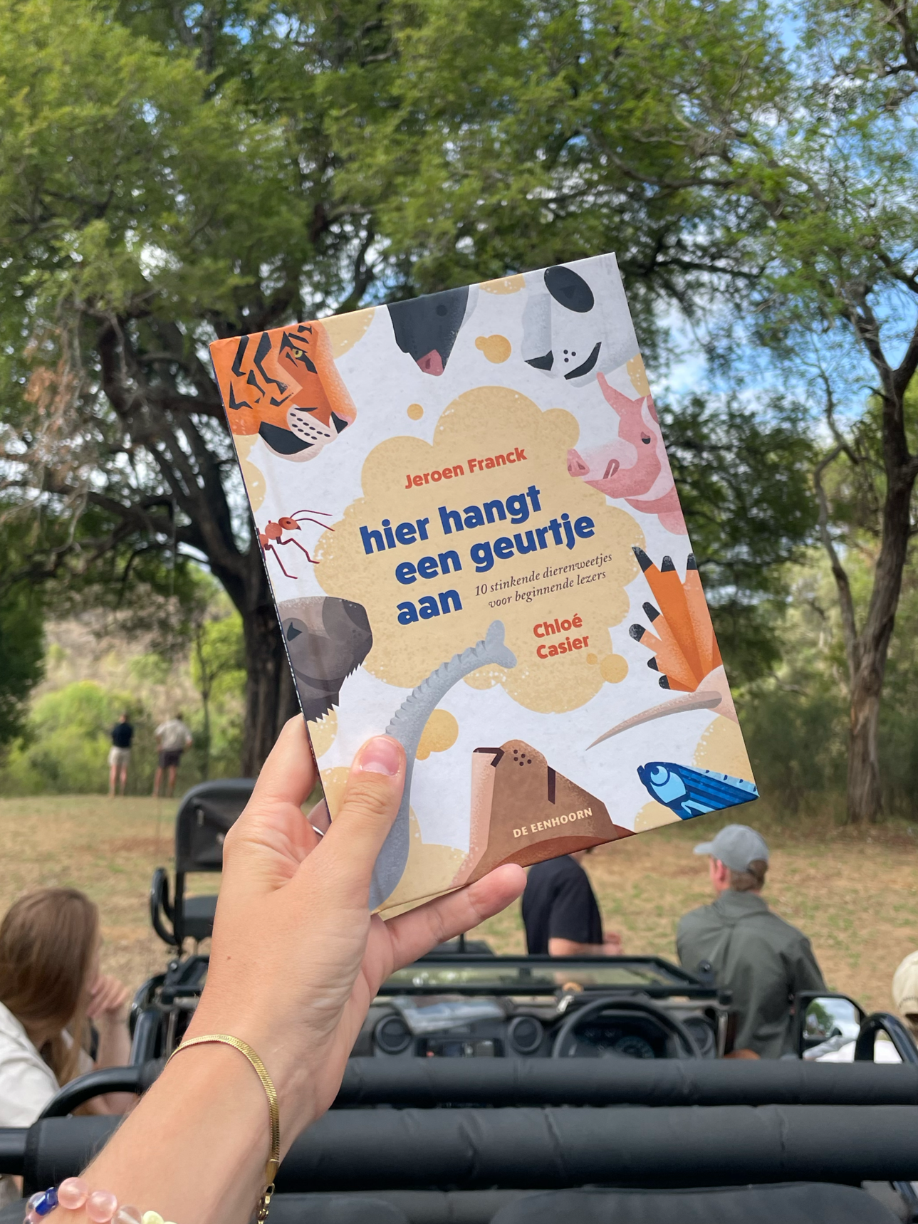A person holds a colorful book titled "hier hangt een geurtje aan" in front of a park scene with trees and people in the background.