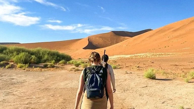 Two people hiking through a desert landscape with large sand dunes and sparse green vegetation under a blue sky.