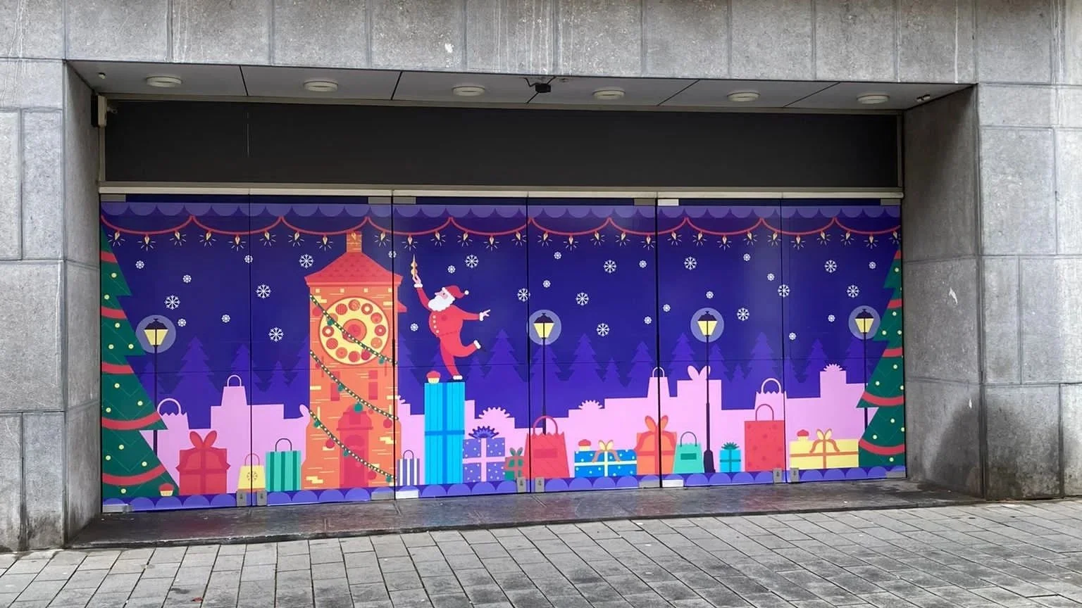 Colorful Christmas-themed artwork on a garage door showing Santa Claus dancing on a tall gift box, surrounded by Christmas trees, decorated presents, street lamps, and falling snow, with a cityscape in the background.