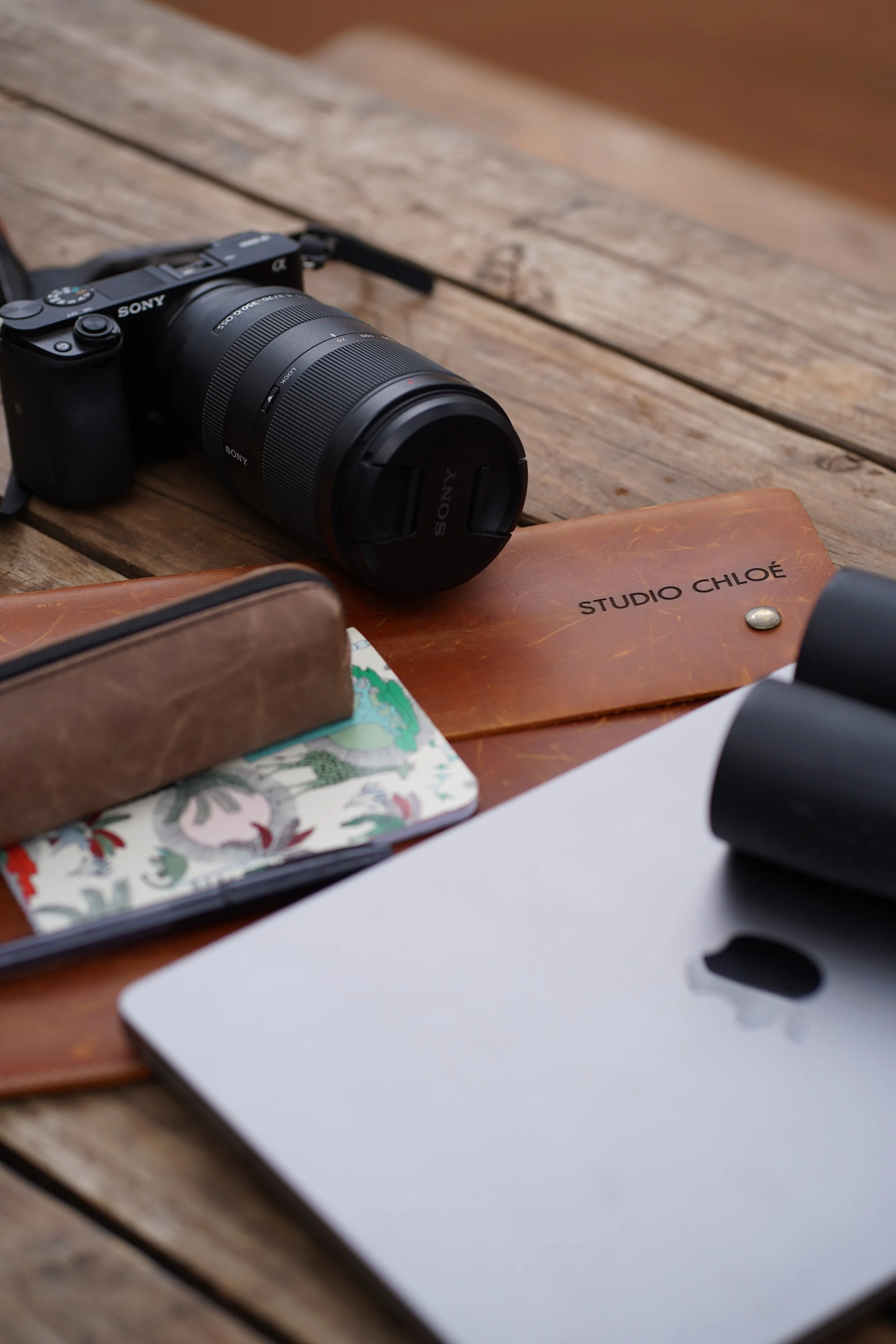 A camera, a leather notebook labeled 'STUDIO CHLOÉ', a laptop, a patterned wallet, and other items scattered on a wooden surface.