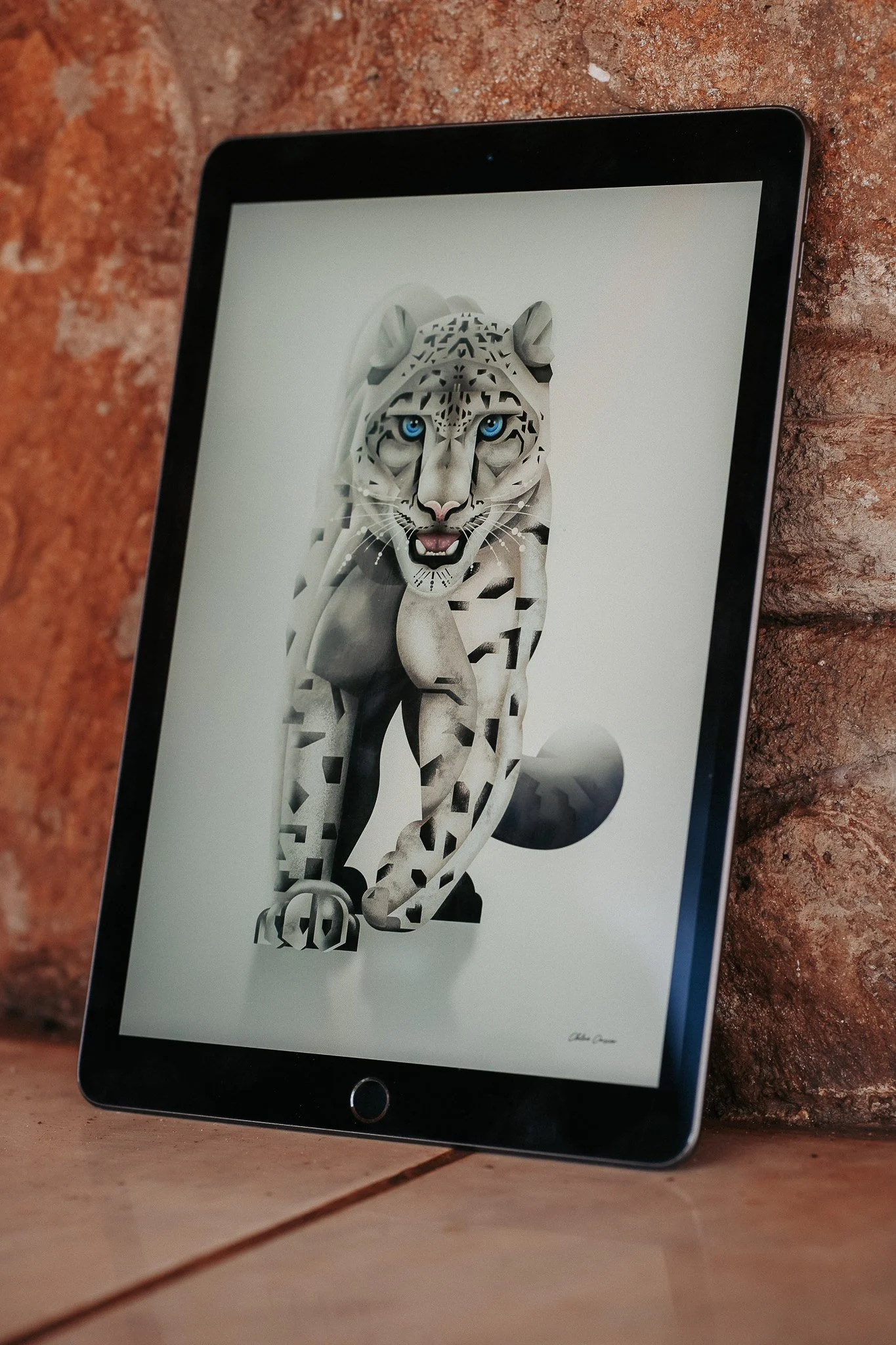 Digital illustration of a white tiger with striking blue eyes, composed of geometric shapes, displayed on a tablet screen resting on a tiled floor by a brick wall.