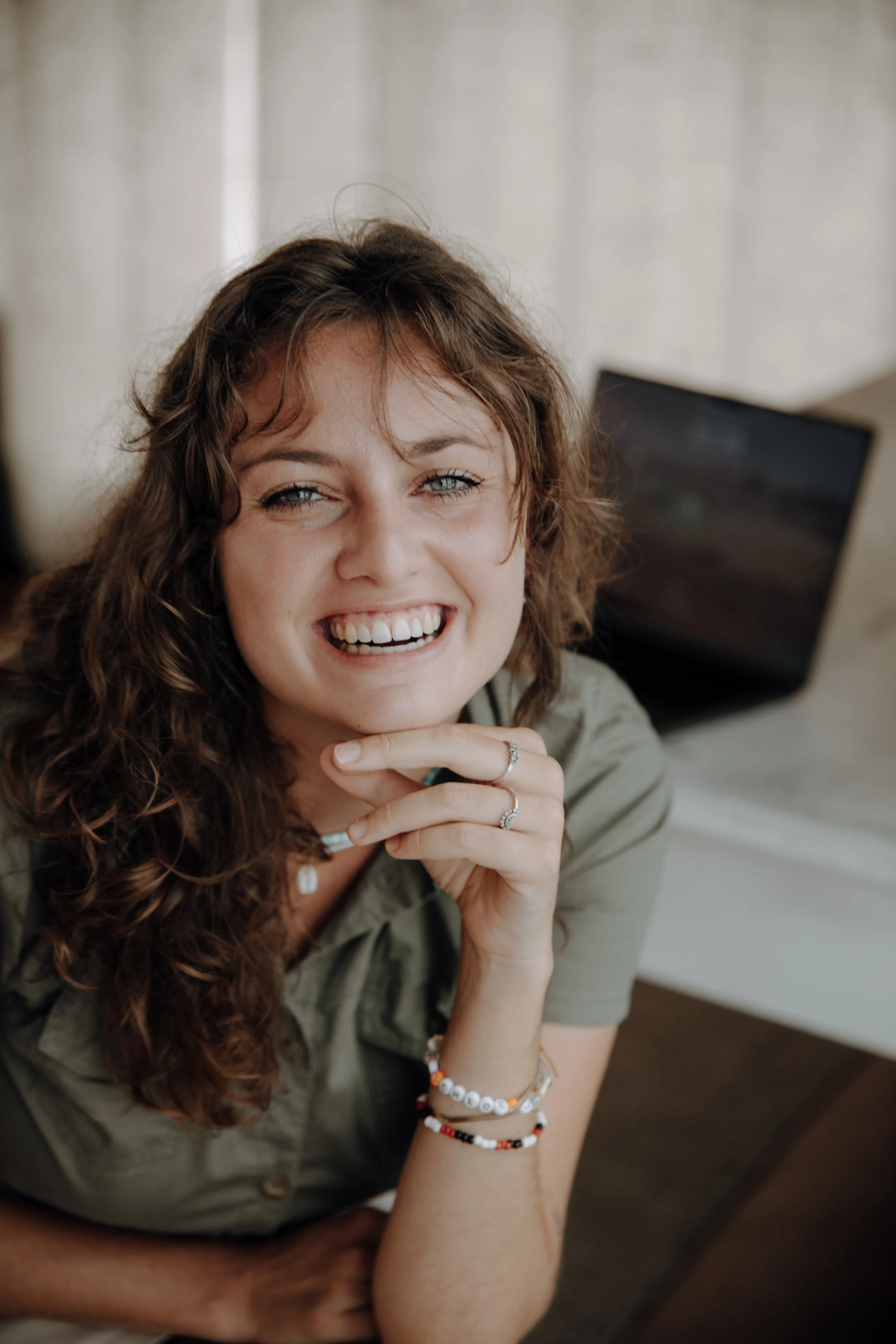 A young woman with curly brown hair smiling and looking at the camera, sitting in front of a laptop on a desk.