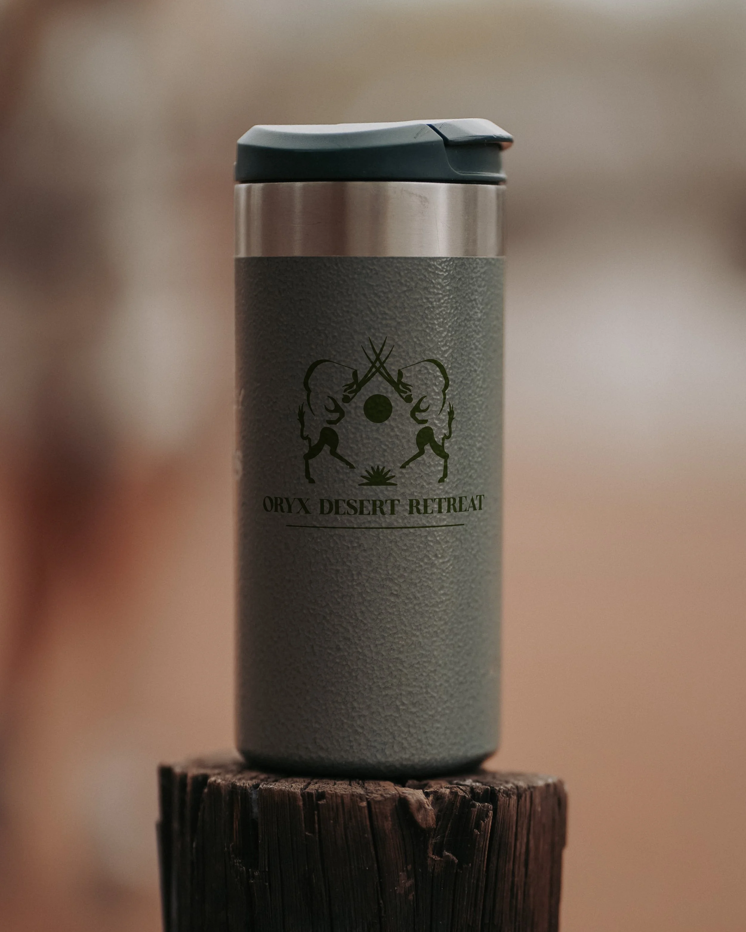 A textured gray travel mug with a black logo and words 'Oryx Desert Retreat' on a wooden post.