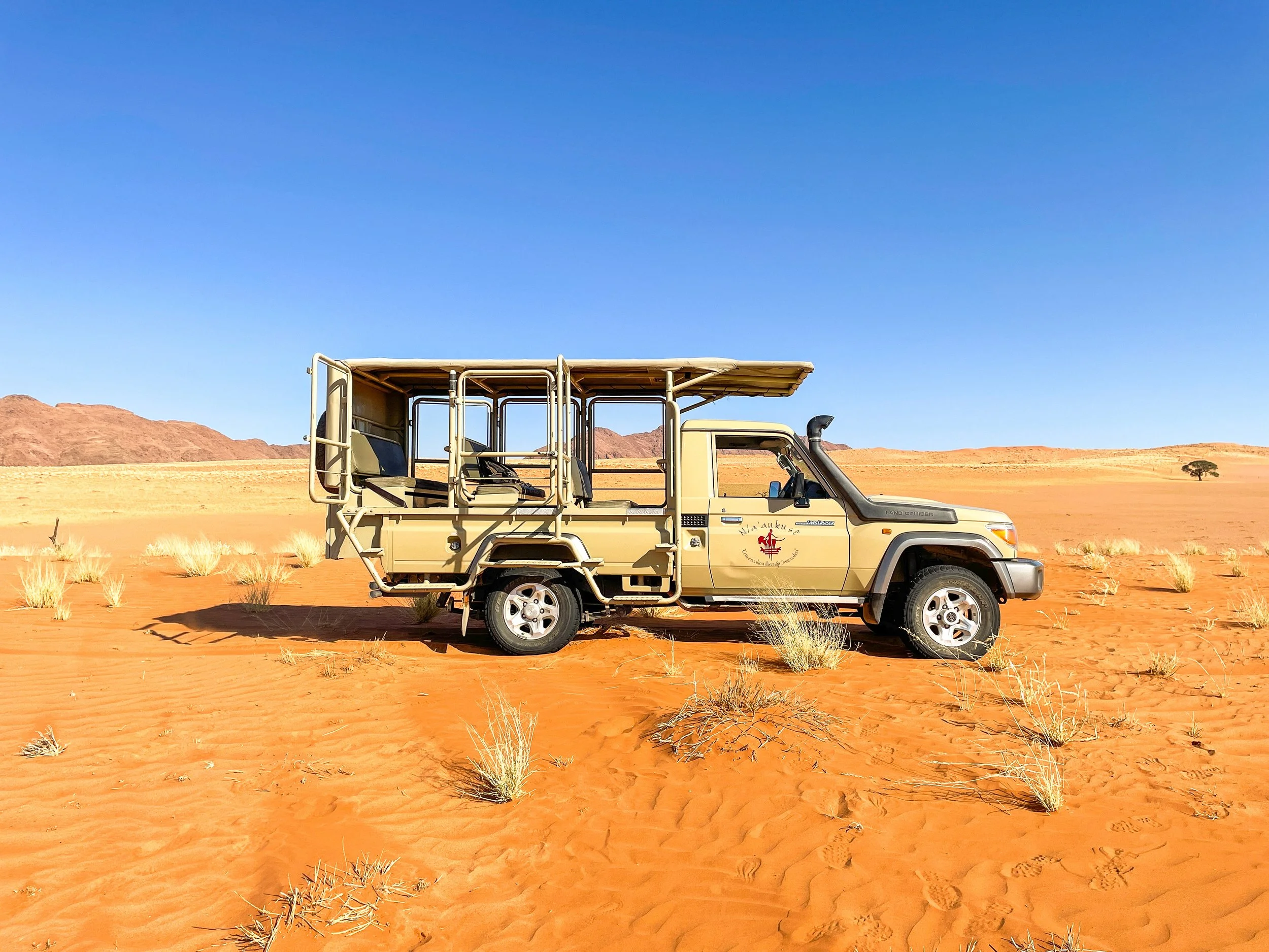 A beige safari vehicle with open sides parked on red desert sand, with sparse bushes and distant mountains under a clear blue sky.