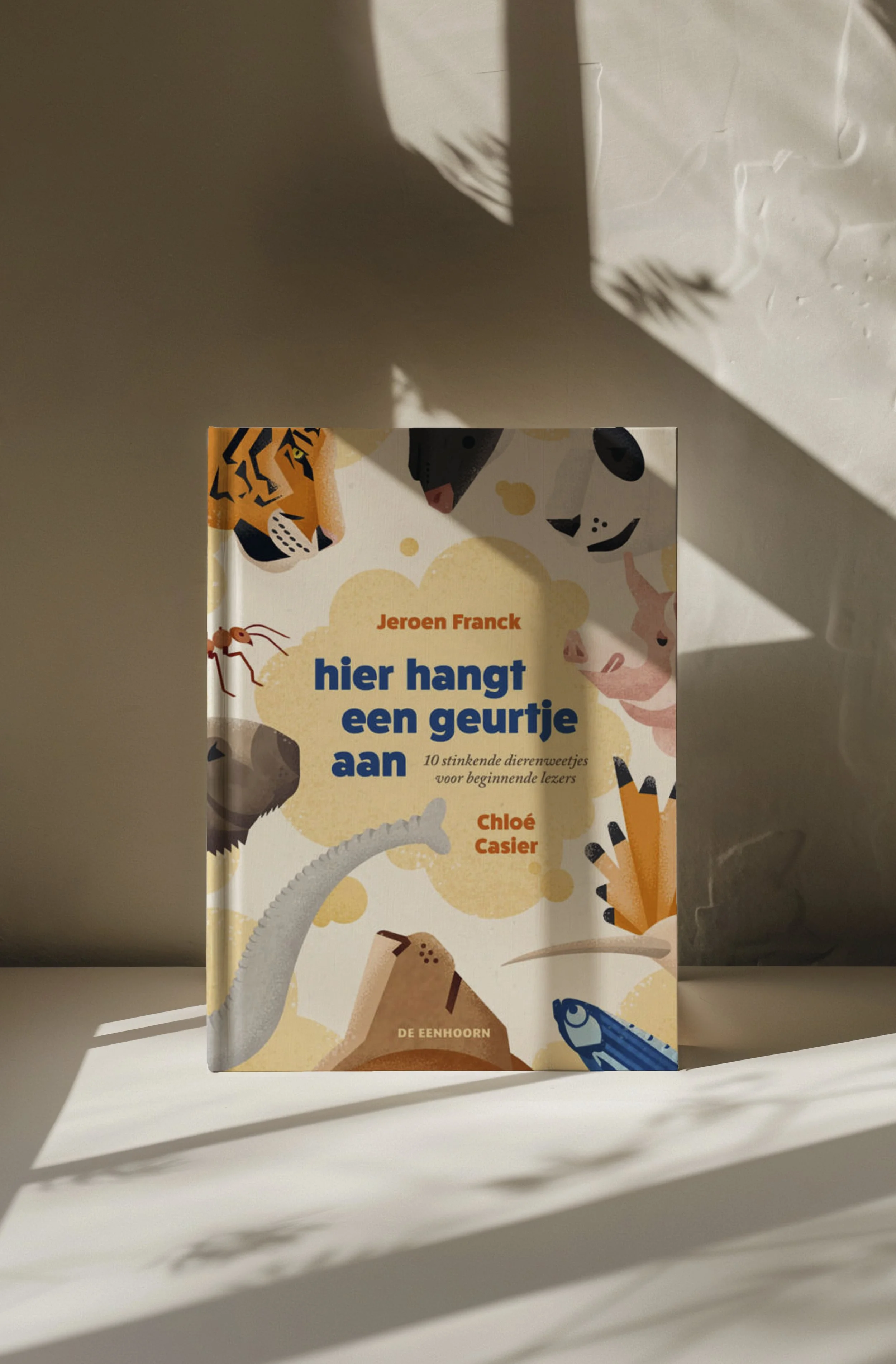 Book titled "Hier hangt een geurtje aan" by Jeroen Franck and Chloé Casier by 'De Eenhoorn' Publisher, featuring colorful illustrations of animals like a tiger, pig, snake, and fish on the cover, placed on a white surface with sunlight and shadows.