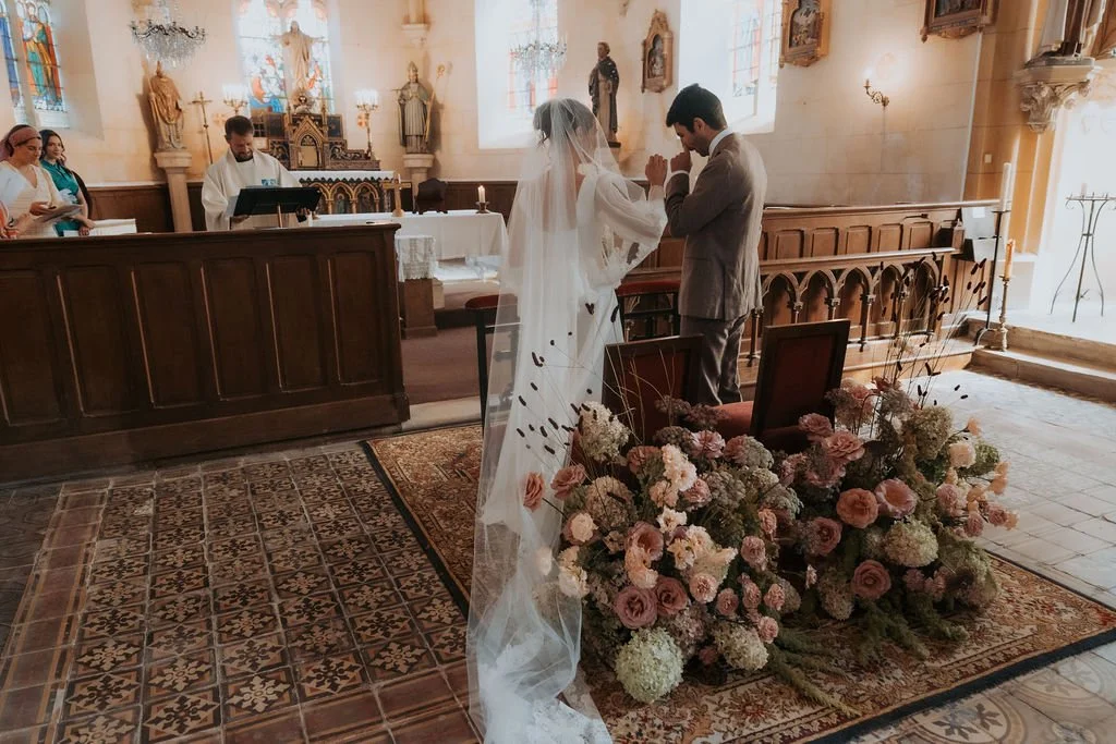 Un couple de mariage priant dans une église, avec un officiaire devant l'autel et des fleurs. Par votre photographe de mariage à paris