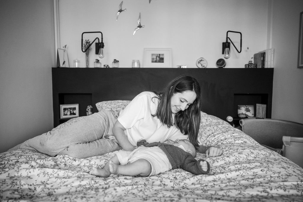 Une femme allongée sur un lit regardant tendrement un bébé couché à côté d'elle dans une chambre décorée sobrement avec des étagères et des objets personnels.