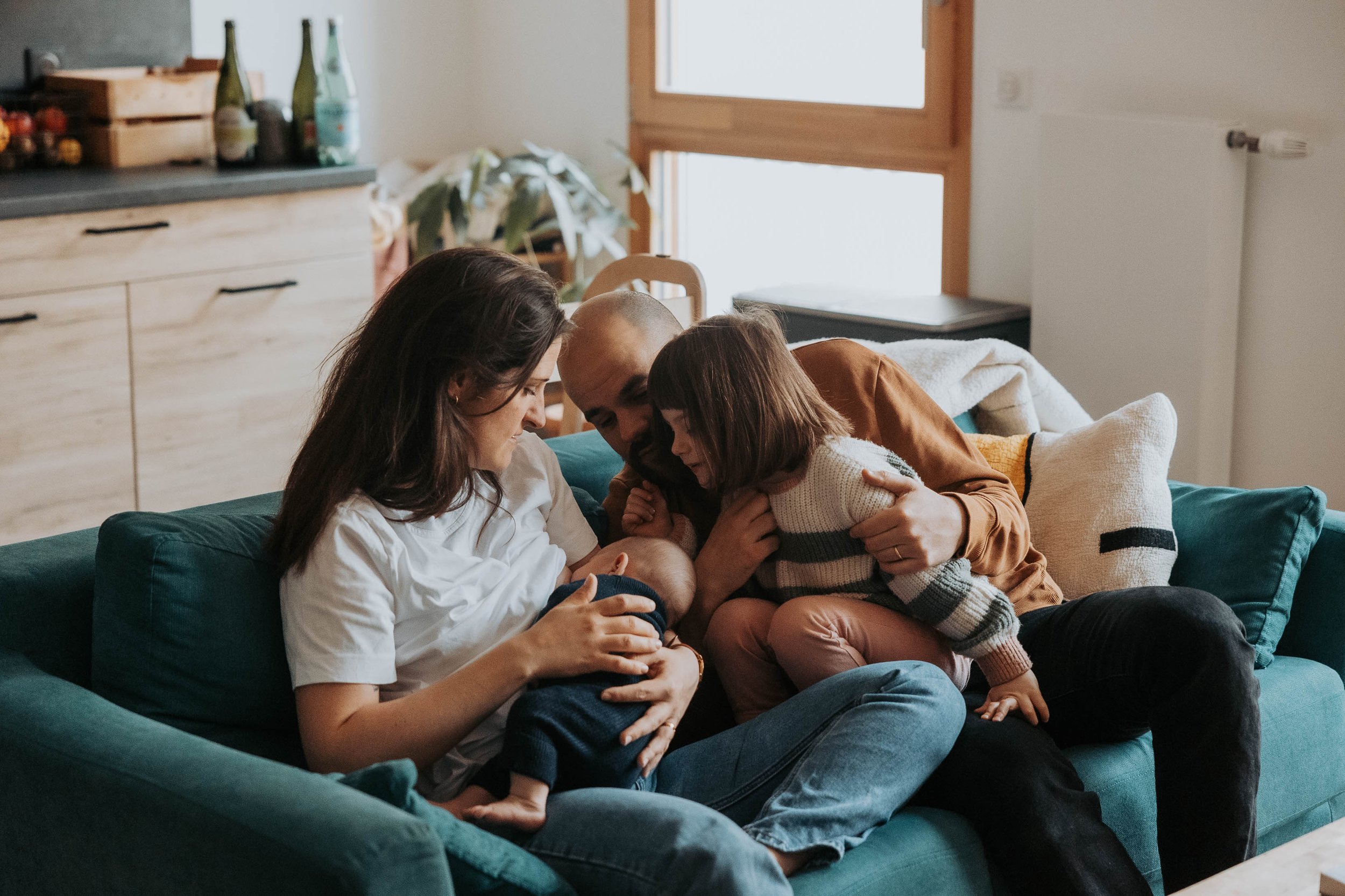 Une famille assise sur un canapé, composée d'un homme, d'une femme, d'une petite fille et d'un bébé. Ils sont dans un salon avec des bouteilles sur une étagère et un mur blanc en arrière-plan.