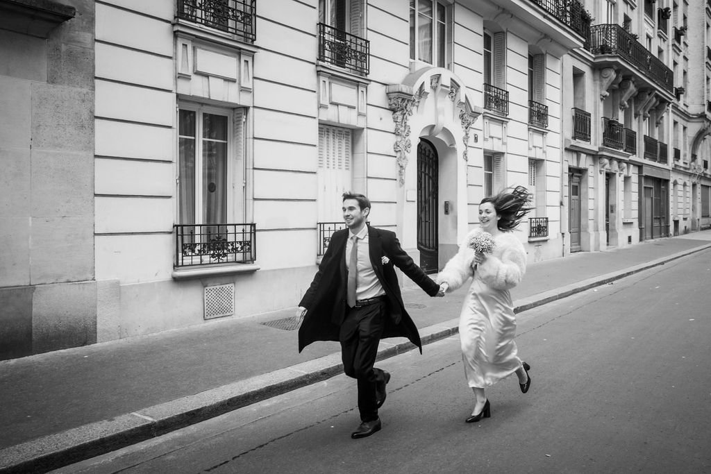 Un couple habillé élégamment court en se tenant la main dans une rue bordée d'immeubles typiquement parisiens. La femme porte une robe blanche et une veste en fourrure avec un bouquet de fleurs. L'homme porte un costume sombre et un manteau long. Ima