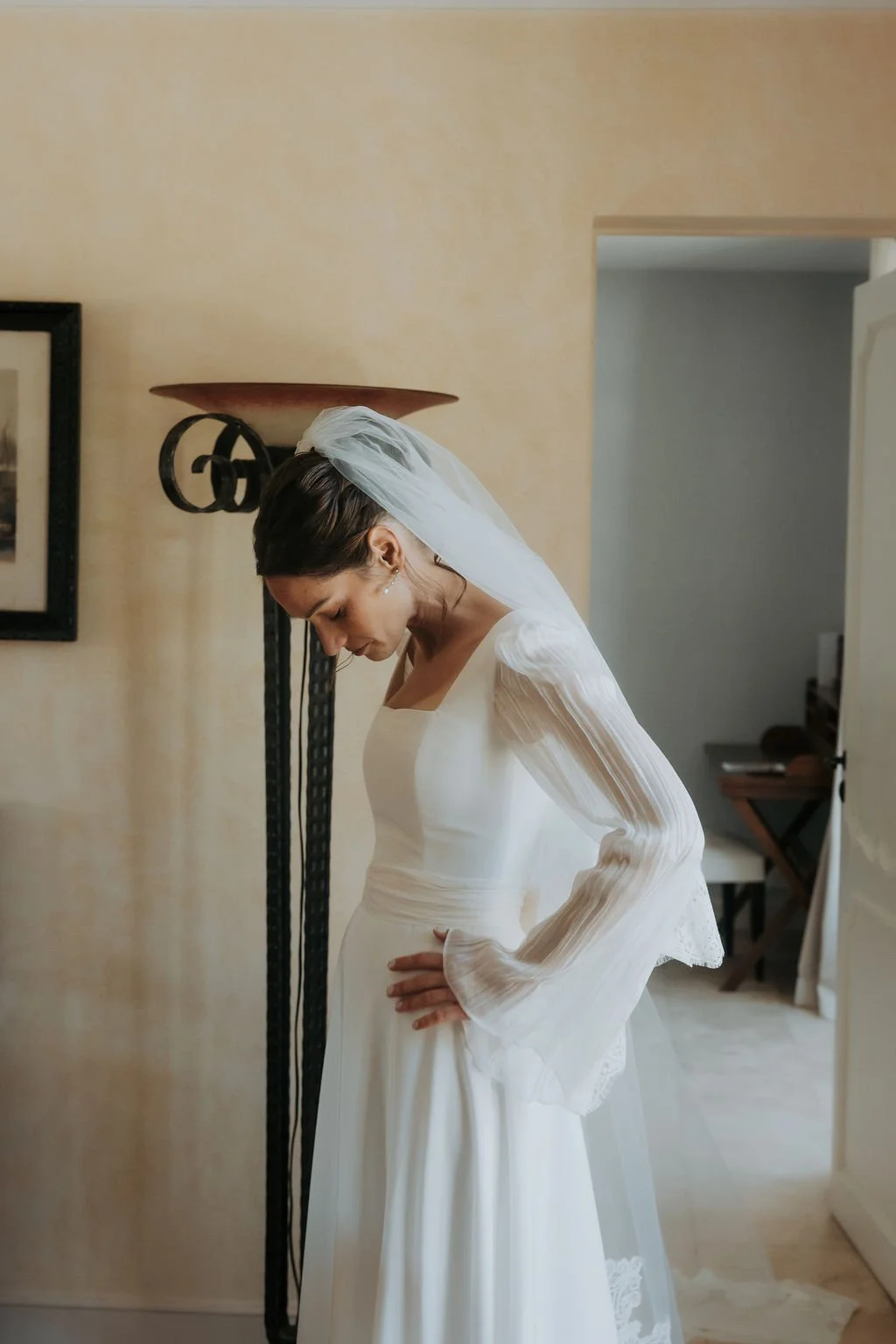 Une femme en robe de mariage blanche, portant un voile, se tient dans une pièce, le regard baissé, les mains sur la taille.