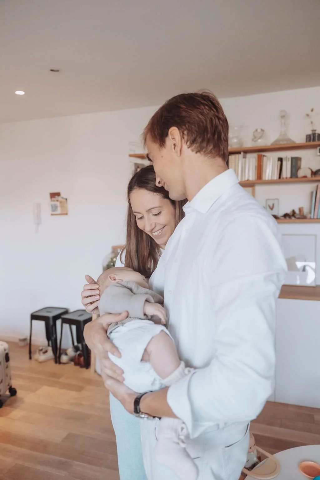Un couple tenant un bébé dans un salon lumineux avec des étagères et des livres en arrière-plan.