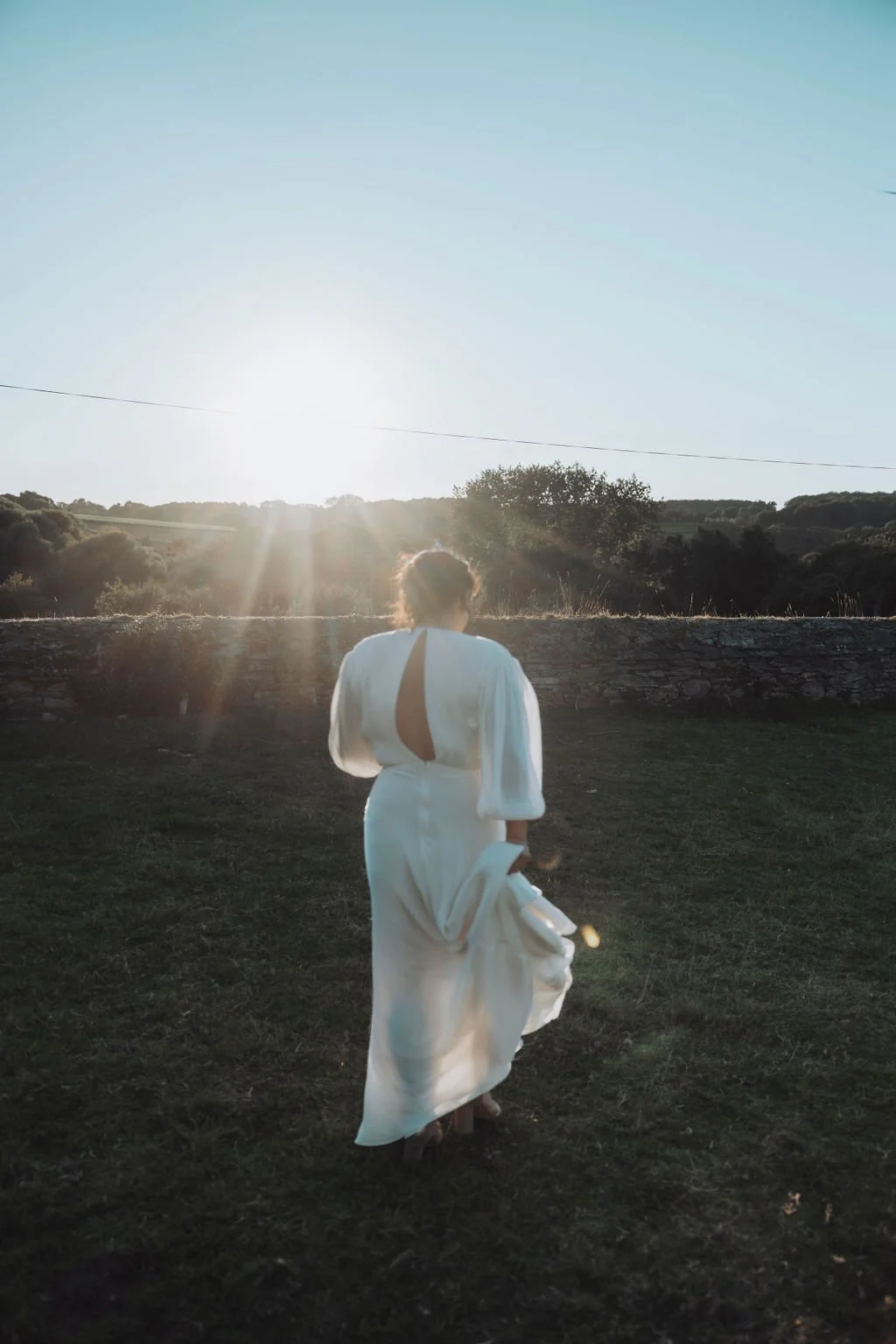 Personne en robe blanche marchant sur une pelouse, avec un coucher de soleil à l'horizon.