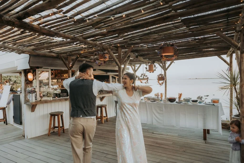 Un couple danse lors d'une réception en plein air sous une toiture en bois, avec un bar et une table à manger en arrière-plan, près de l'eau, un enfant à droite. Mariage en bretagne par une photographe de mariage