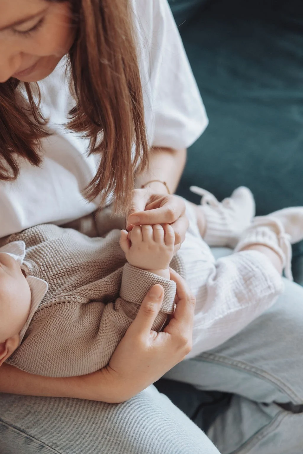 Une femme tient et regarde tendrement un bébé habillé de vêtements en laine beige, assis sur ses genoux. Le bébé porte des chaussons blancs et tient la main de la femme.