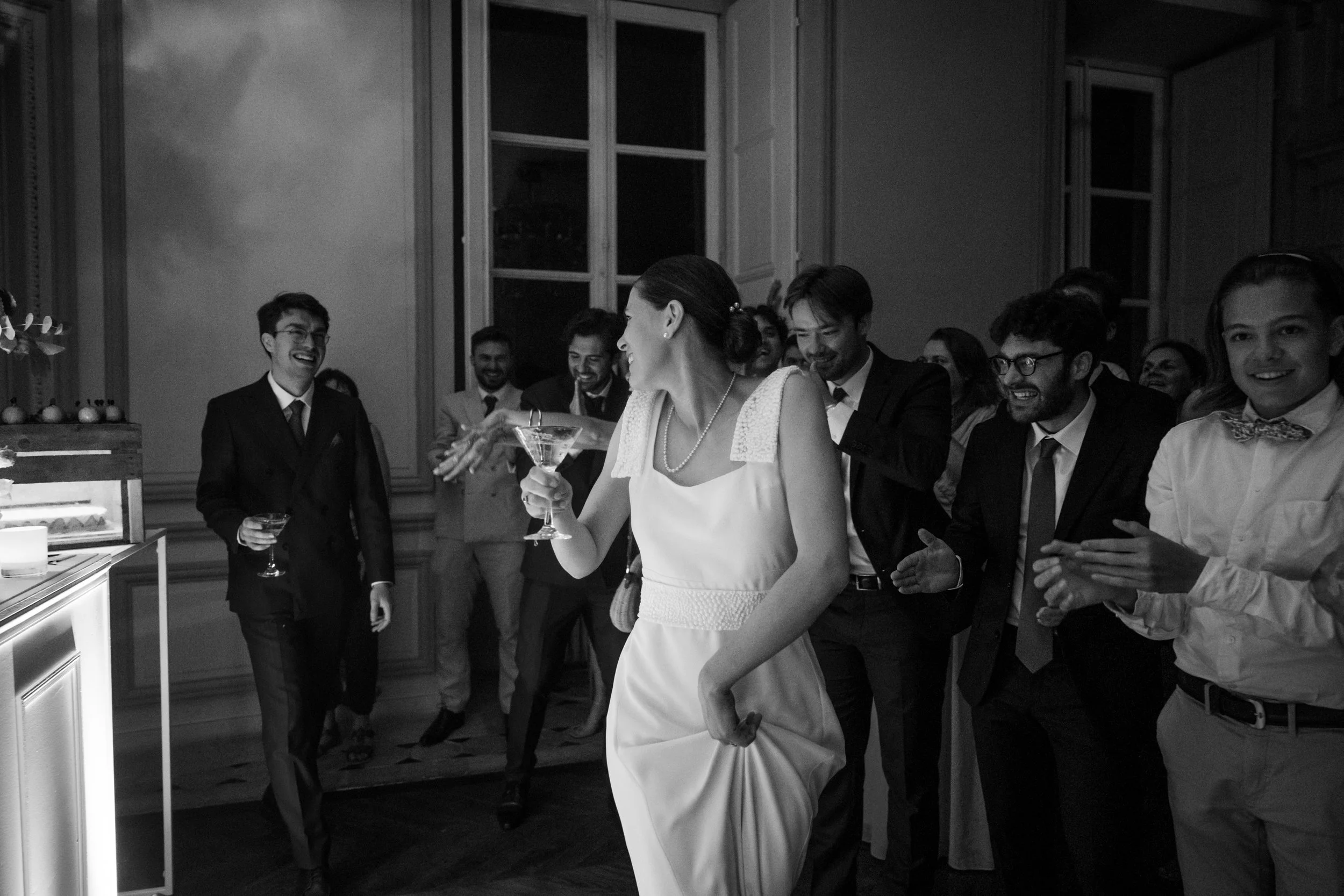 Groupe de personnes lors d'une fête de mariage, avec une femme au centre tenant un verre, souriante et entourée d'hommes habillés en costumes, dans une pièce élégante.