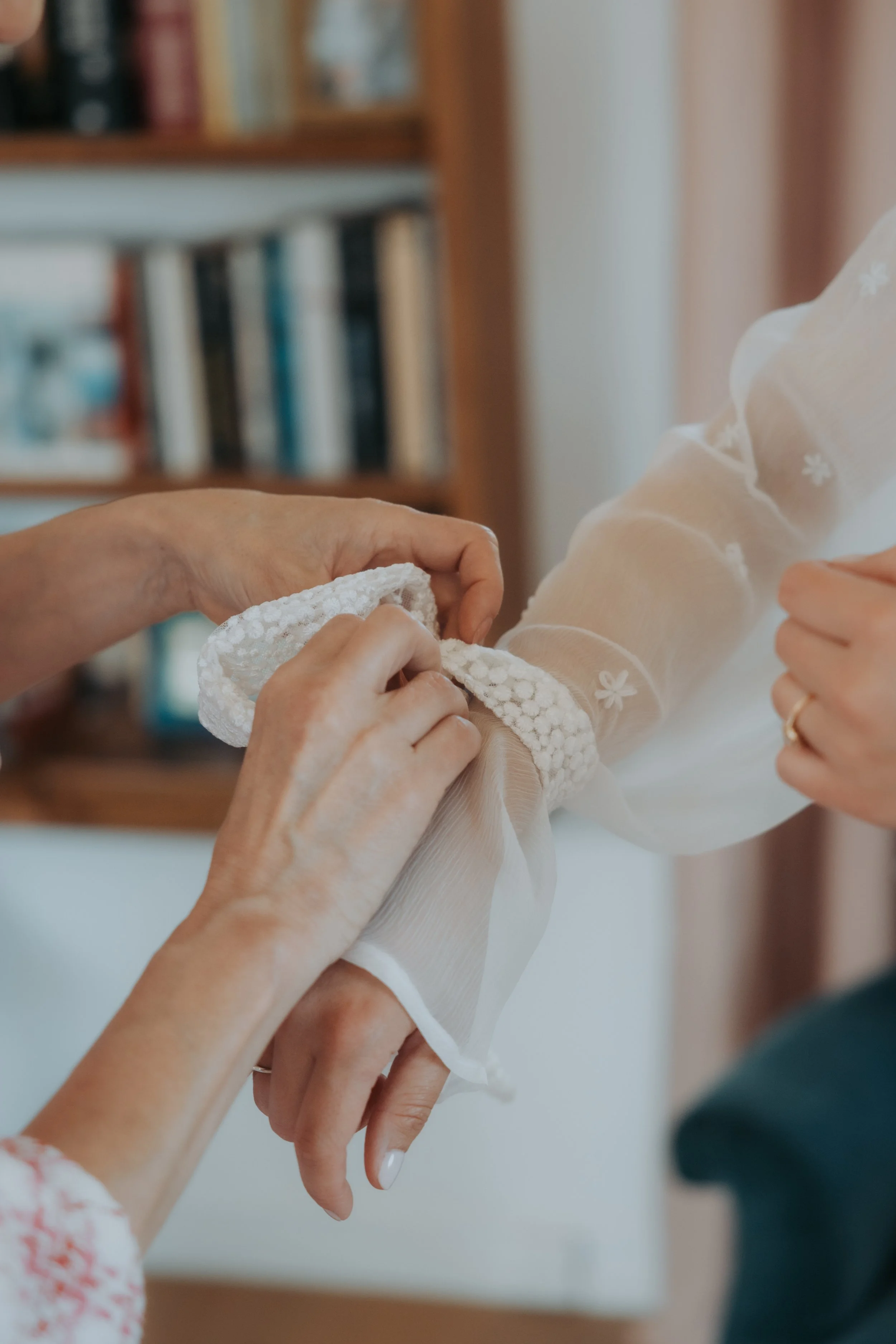 Une personne attache un bracelet en tissu blanc avec des fleurs à une autre personne portant une chemise en voile transparent avec des broderies. En arrière-plan, une étagère avec des livres. photo de mariage