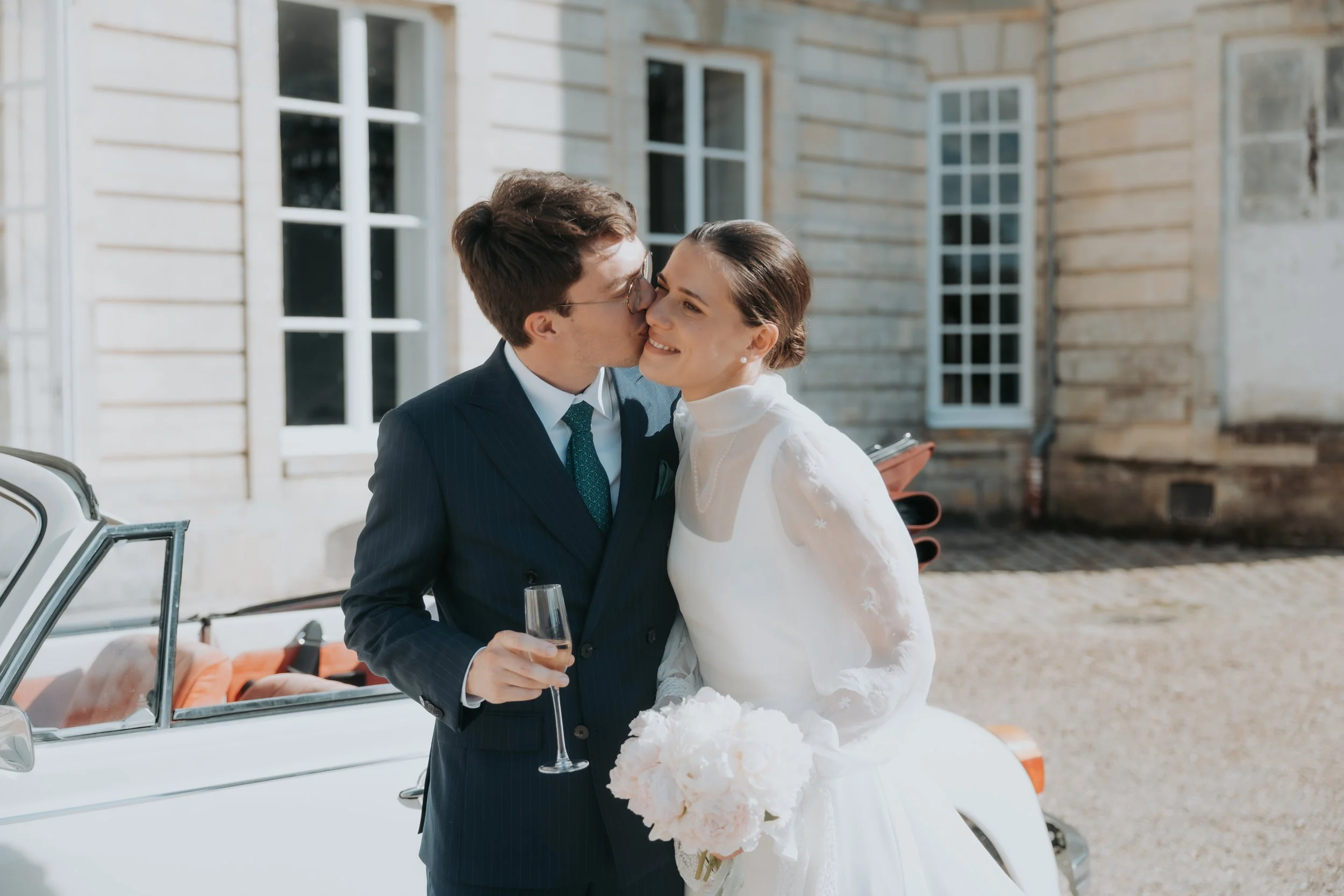 Un couple de mariés s'embrasse avec un sourire en tenant un bouquet de fleurs blanches, devant une voiture classique, à l'extérieur d'une bâtisse en pierre.