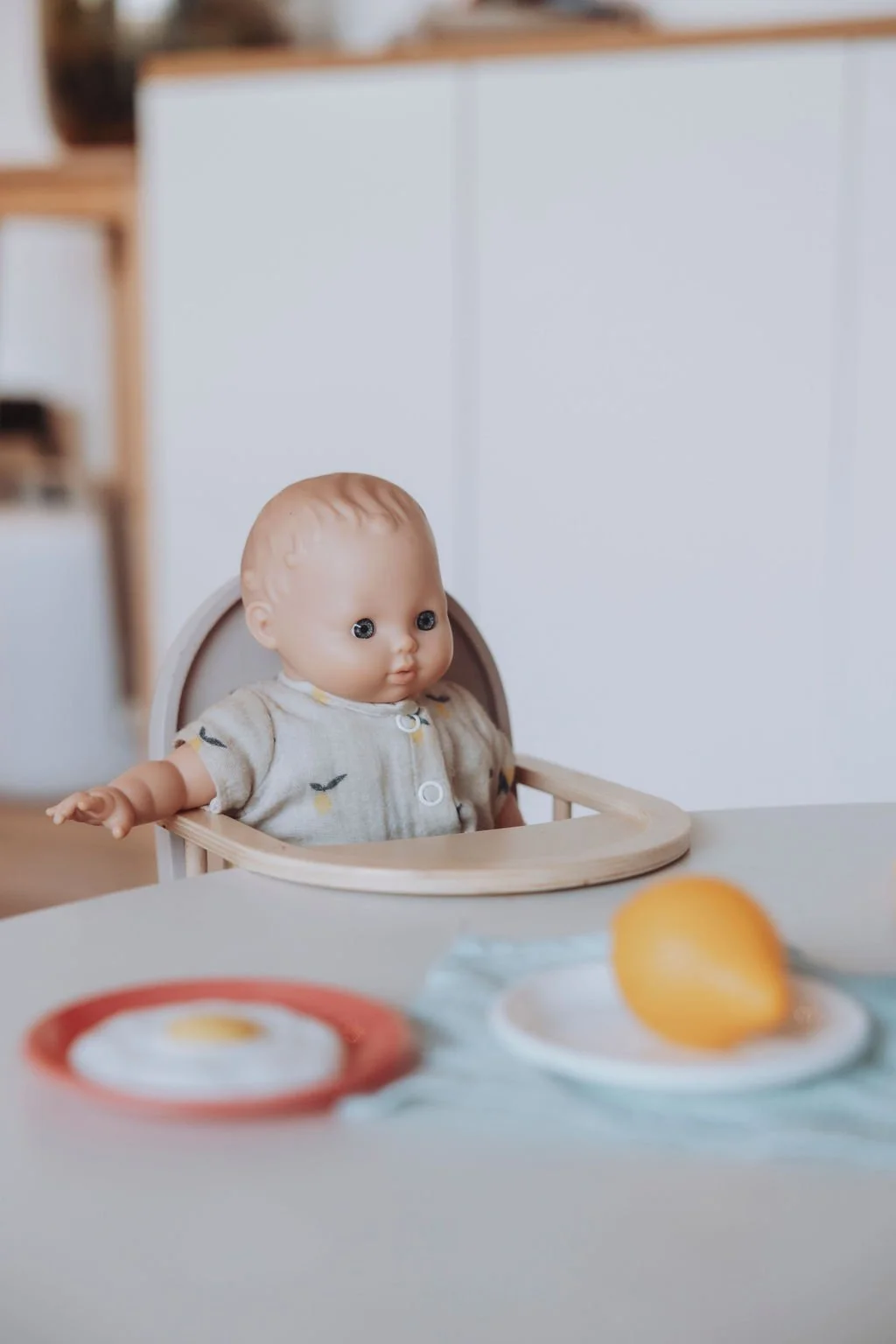 Un poupon assis dans une chaise haute avec une assiette contenant un œuf au plat en plastique et un citron en plastique sur une table.