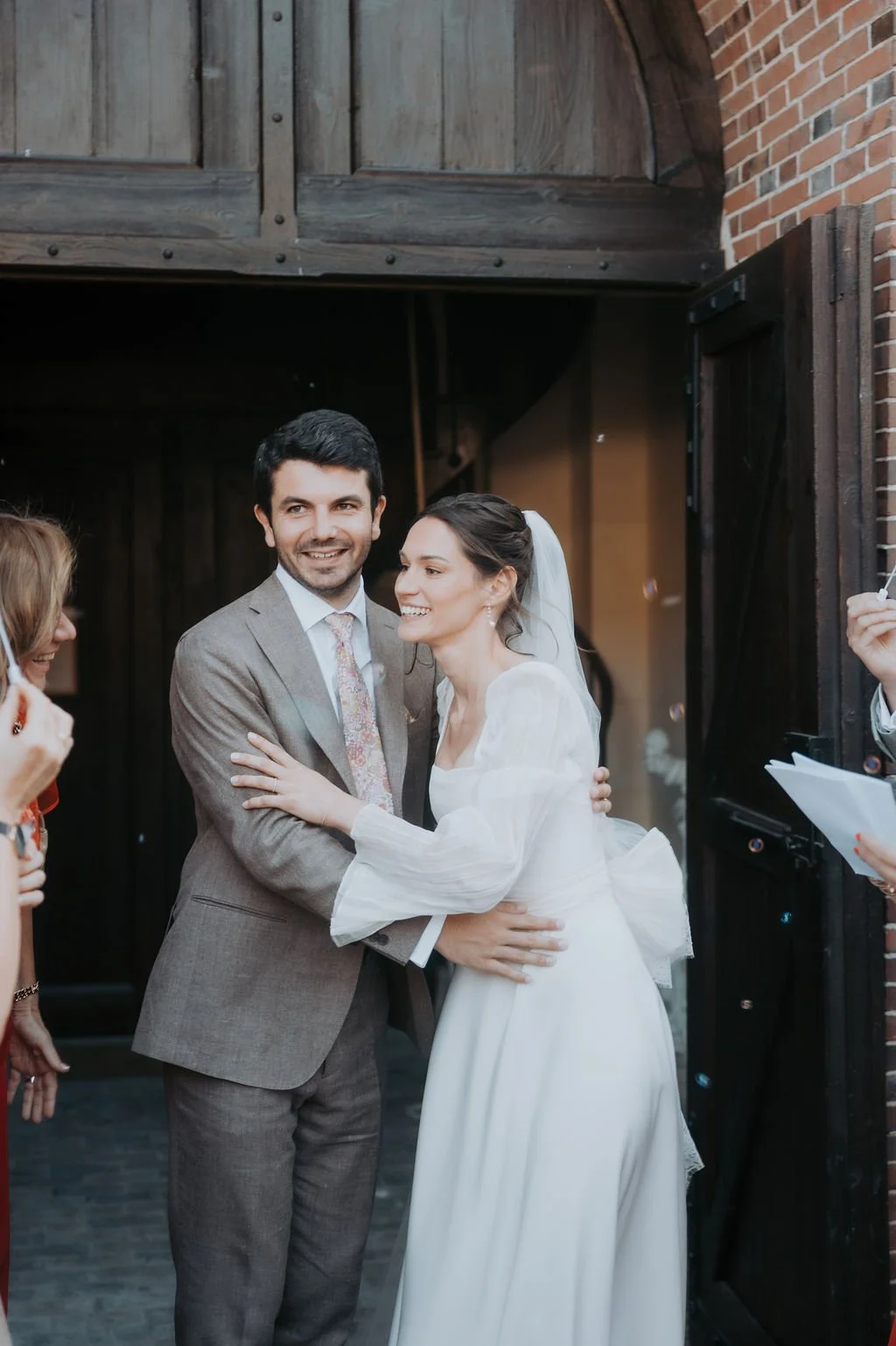 Un couple de mariés souriants, l'un en costume gris et l'autre en robe blanche, partage un moment heureux à leur mariage, entourés d'invités. Par votre photographe de mariage à paris