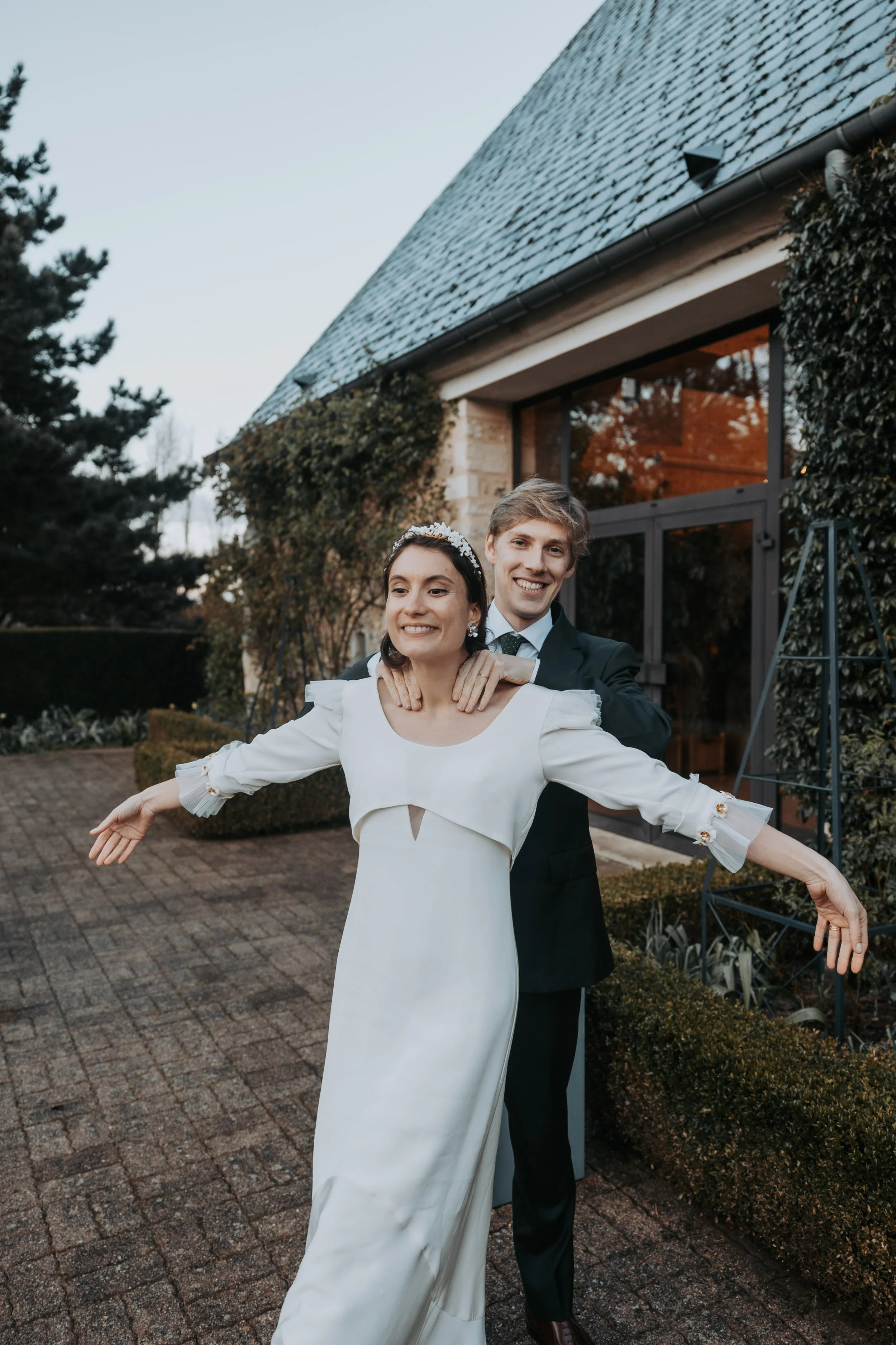 Un couple souriant célébrant leur mariage devant un bâtiment en pierre avec des vignes grimpantes.