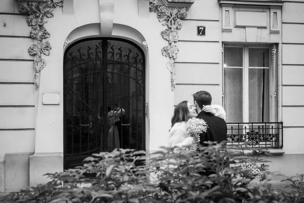 Couple embrassant devant un bâtiment ancien, photographié en noir et blanc avec des plantes en premier plan.