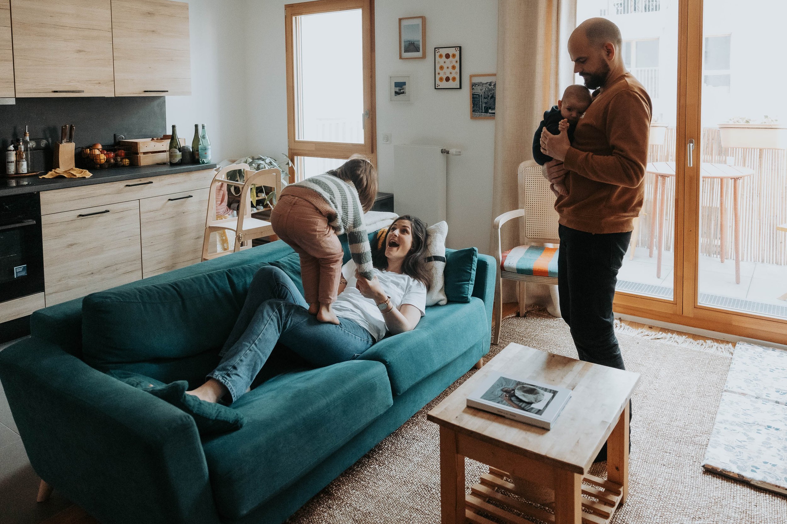 Une famille dans un salon lumineux, une femme allongée sur le canapé vert joue avec un enfant debout sur elle, pendant qu'un homme tient un bébé près de la fenêtre. Une cuisine moderne se trouve à l'arrière-plan.