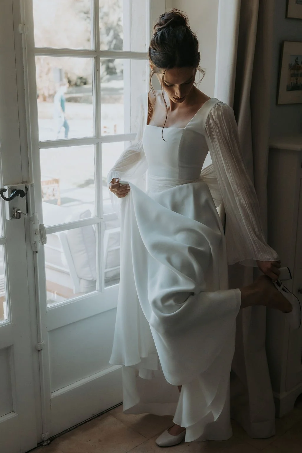 Une femme en robe blanche de mariage se prépare à marcher, ajustant sa chaussure en haut d'un escalier près d'une porte-fenêtre.