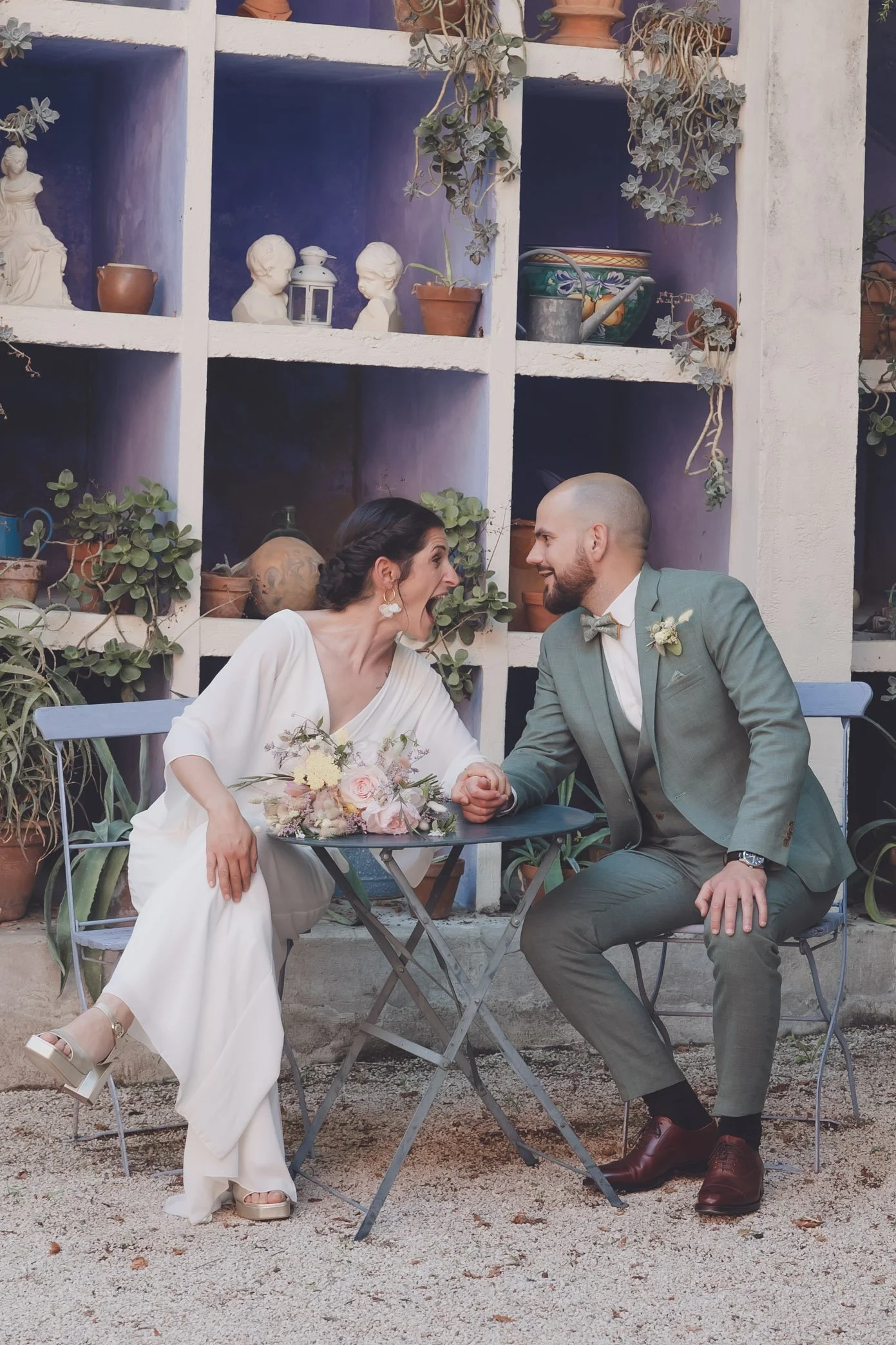 Un couple assis à une petite table en plein air, elle en robe blanche avec un bouquet de fleurs et lui en costume vert. Ils se regardent avec complicité, entourés de plantes et de pots décoratifs.