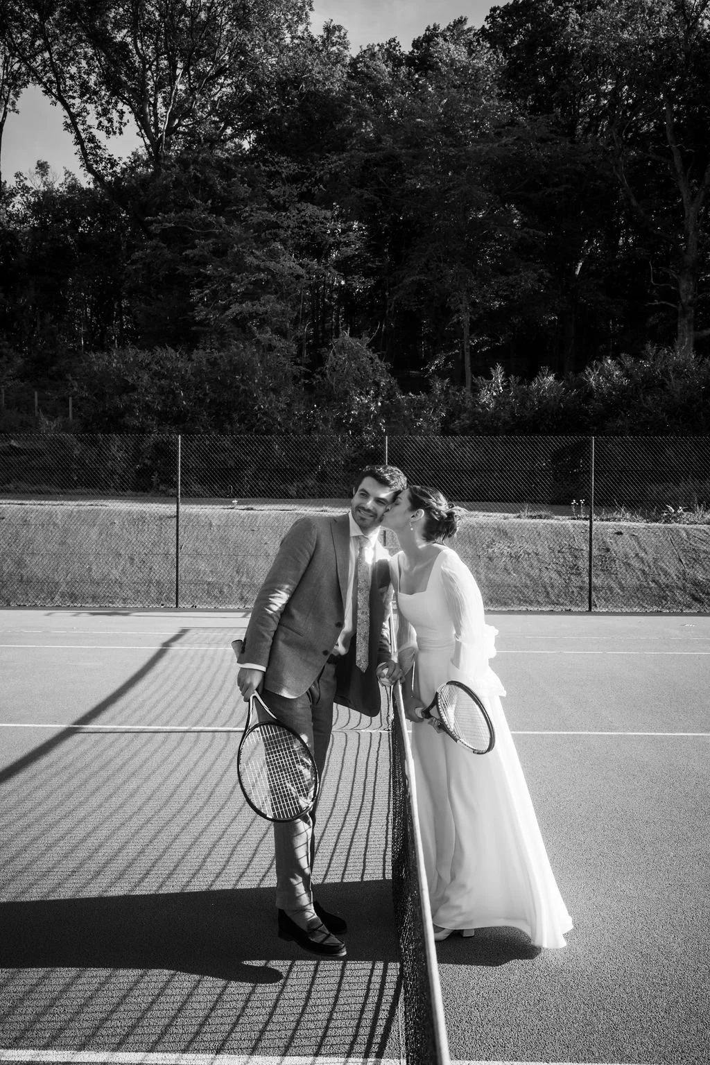 Un couple en tenue de mariage jouant au tennis sur un court en extérieur, la femme embrassant l'homme.