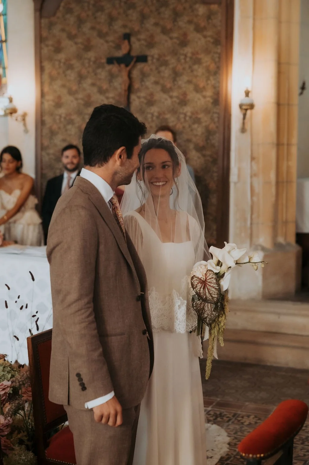 Un couple de mariage échangeant leurs vœux dans une église avec un fond en pierre et une croix en haut, entouré de témoins et invités. Par votre photographe de mariage à paris