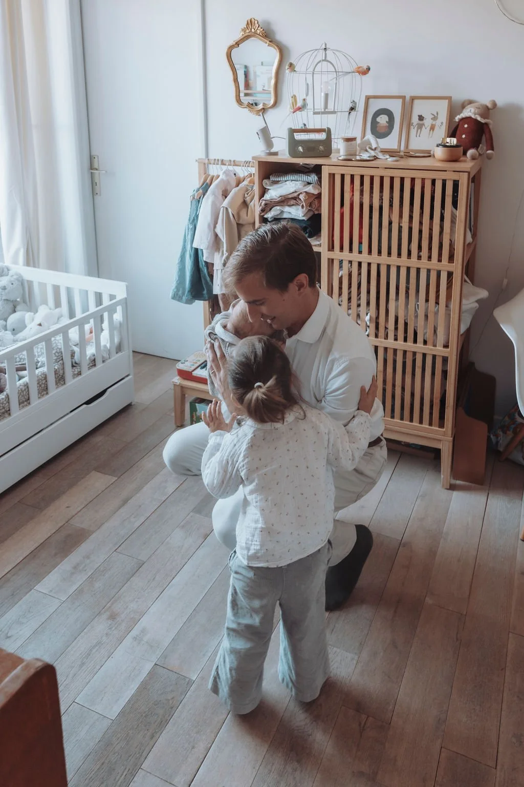 Une pièce avec un homme serrant une petite fille dans ses bras. Un berceau blanc est visible à gauche, et une étagère en bois avec des vêtements et des objets décoratifs se trouve à droite. Un miroir doré est suspendu au mur au-dessus de l'étagère.