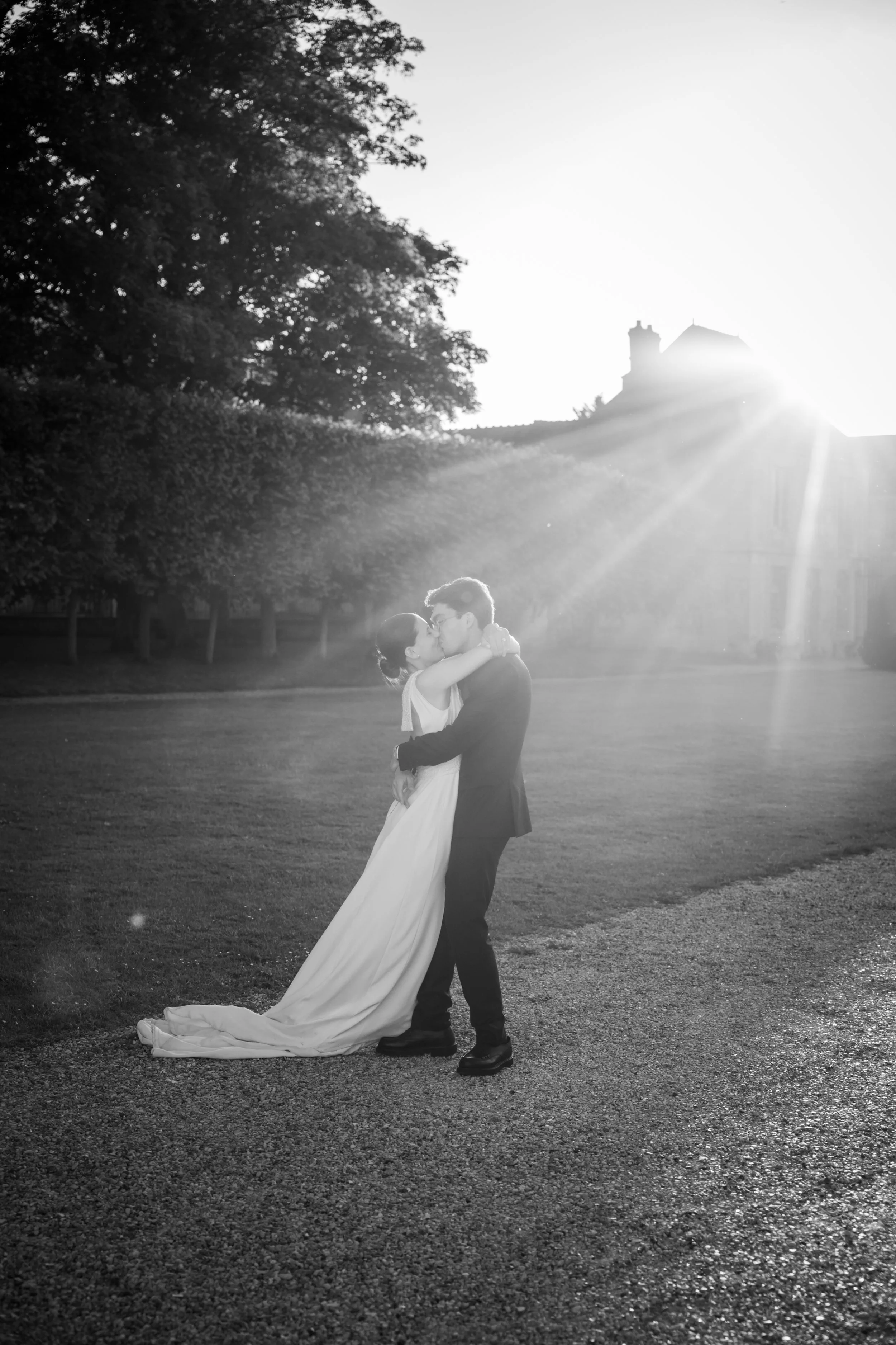 Un couple de mariés s'embrasse en plein air lors d'un coucher de soleil, en noir et blanc, avec des arbres en arrière-plan et une lumière solaire intense derrière eux. Photo de mariage à paris