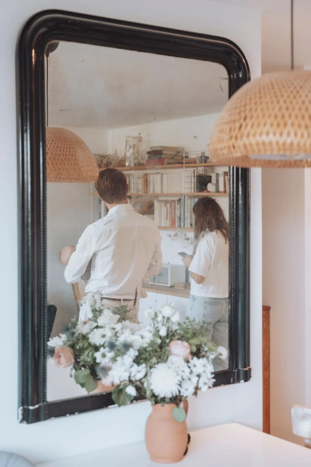 Un miroir reflète un homme tenant un bébé et une femme utilisant un téléphone dans une pièce avec des étagères remplies de livres. Au premier plan, un vase de fleurs est visible.