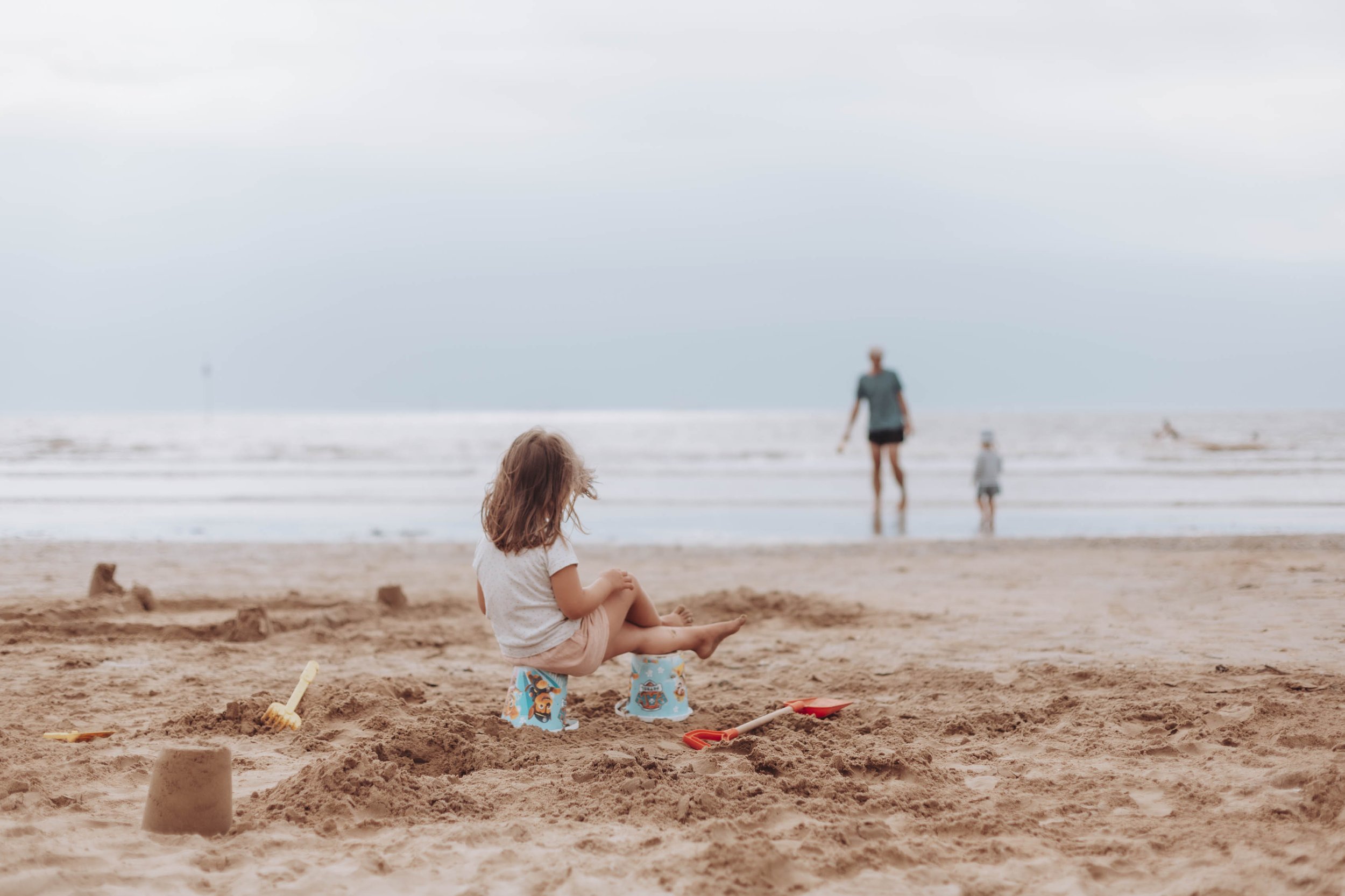 Enfant assis sur un seau en plastique sur une plage, jouant avec des jouets de sable, avec une mer à l'arrière-plan et des personnes marchant dans l'eau.