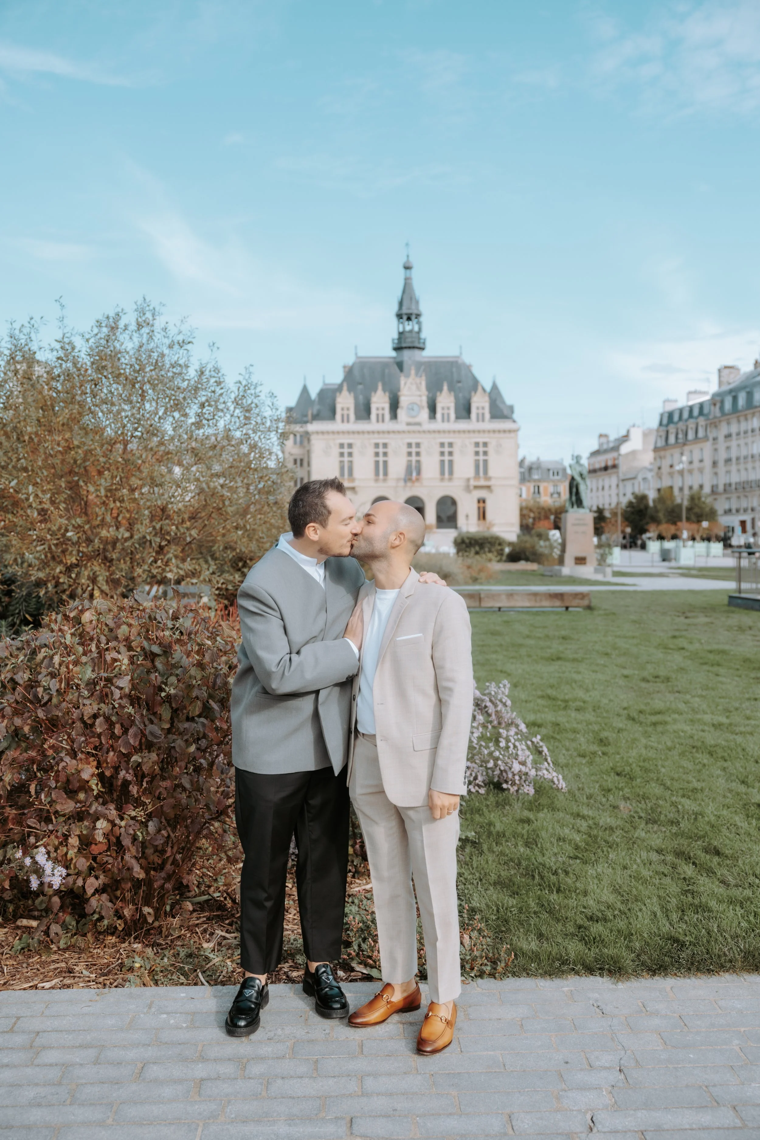 Deux hommes s'embrassent dans un parc en ville, avec un bâtiment historique en arrière-plan. Par votre photographe de mariage à paris