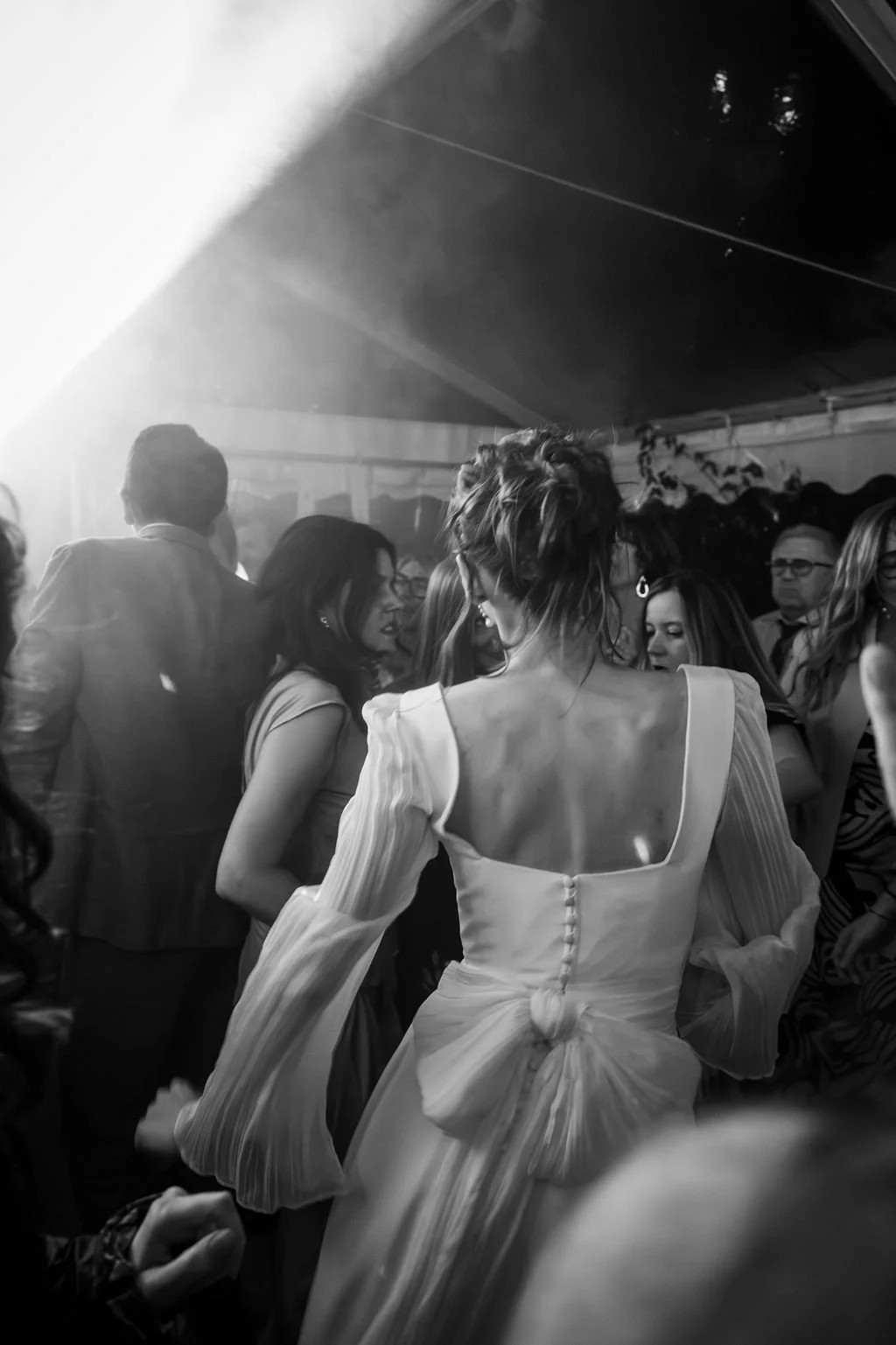 Une femme en robe blanche élégante avec un nœud dans le dos, danse lors d'une fête ou un mariage, entourée d'autres personnes dans une ambiance festive.