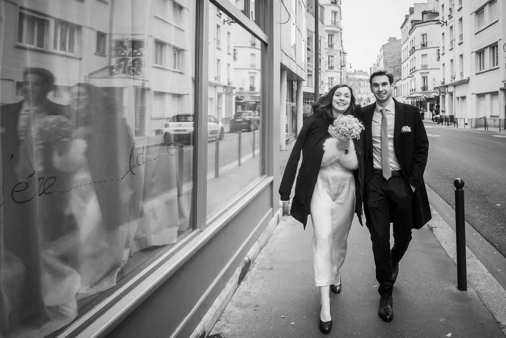 Un couple habillé élégamment marche dans la rue, souriant et tenant un bouquet de fleurs. Leur reflet apparaît dans la vitre d'un bâtiment à côté d'eux.