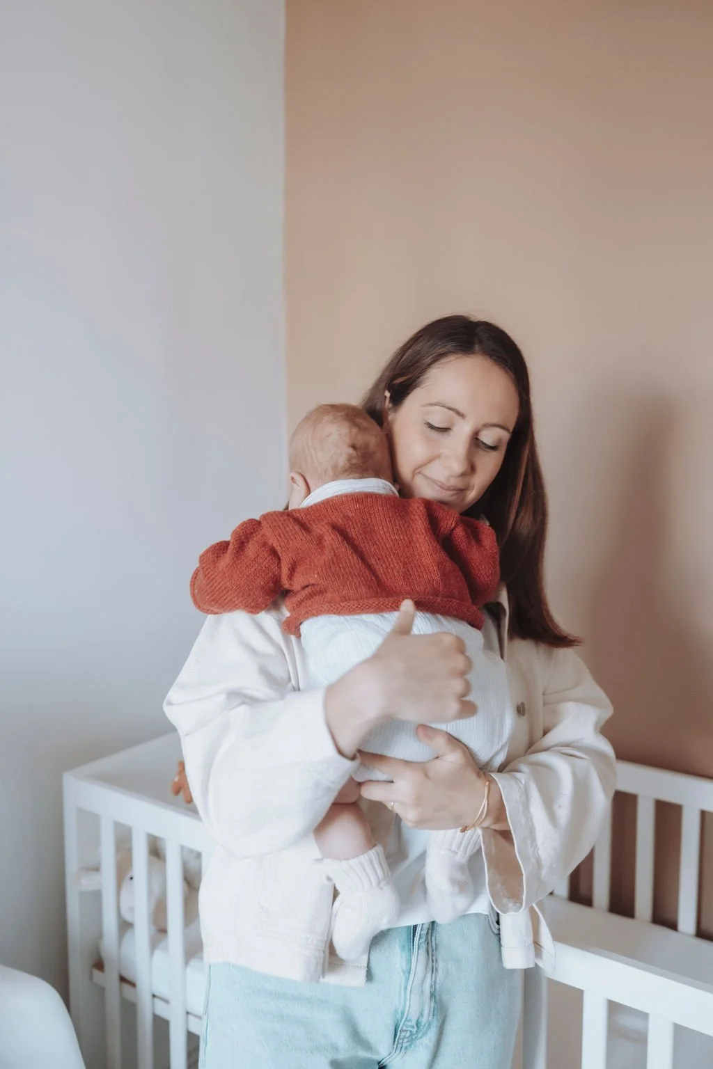 Une femme tient un bébé dans ses bras devant un berceau, avec un mur beige en arrière-plan.
