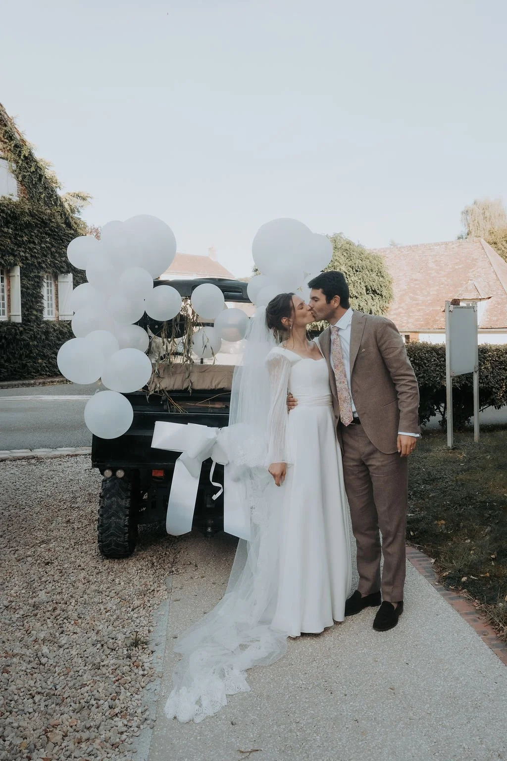 Un couple de mariés s'embrasse devant une remorque décorée de ballons blancs lors d'un mariage en extérieur.