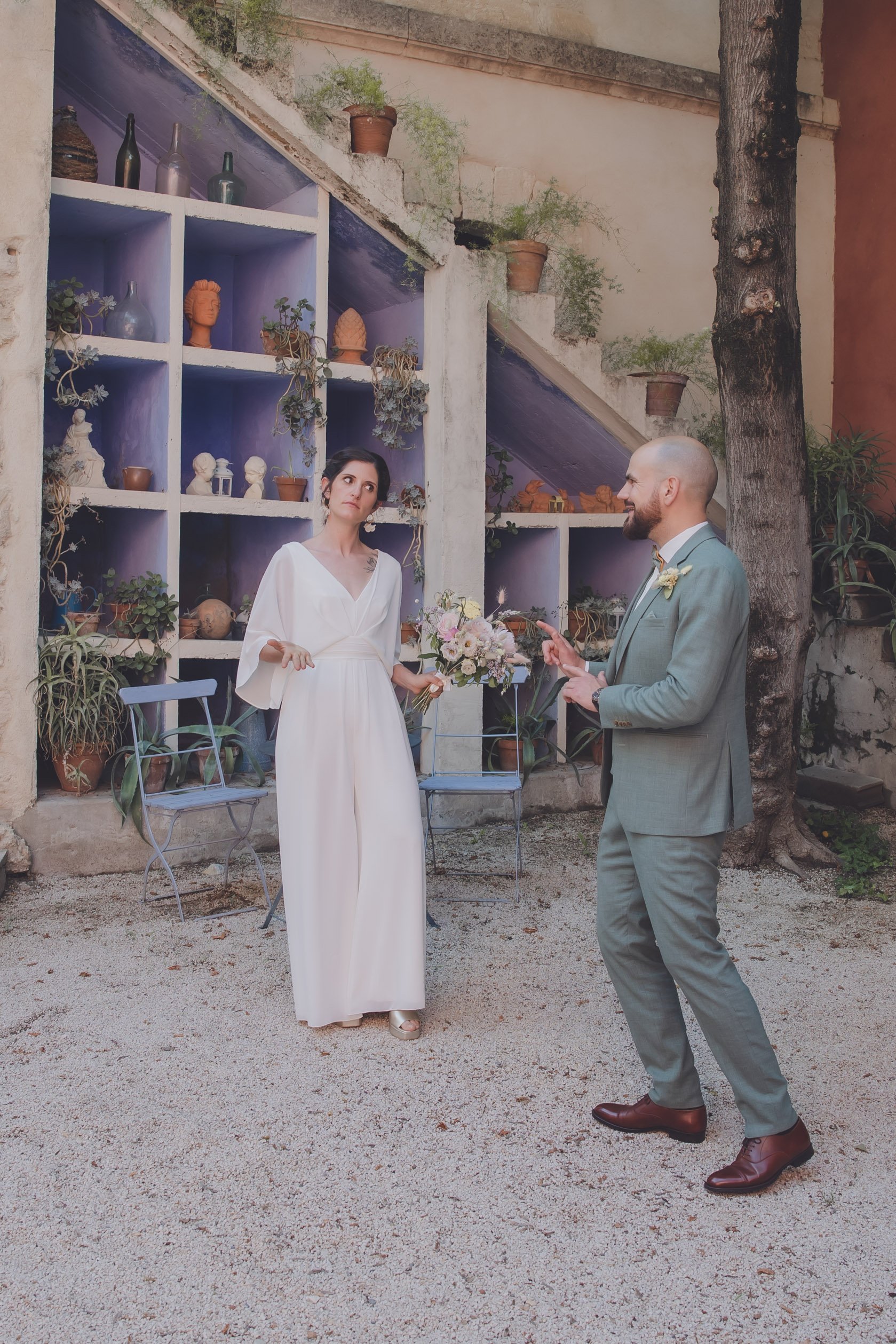 Un couple se tenant debout dans un jardin. La femme porte une robe blanche et tient un bouquet de fleurs. L'homme porte un costume gris. En arrière-plan, une étagère avec des plantes et des objets décoratifs.