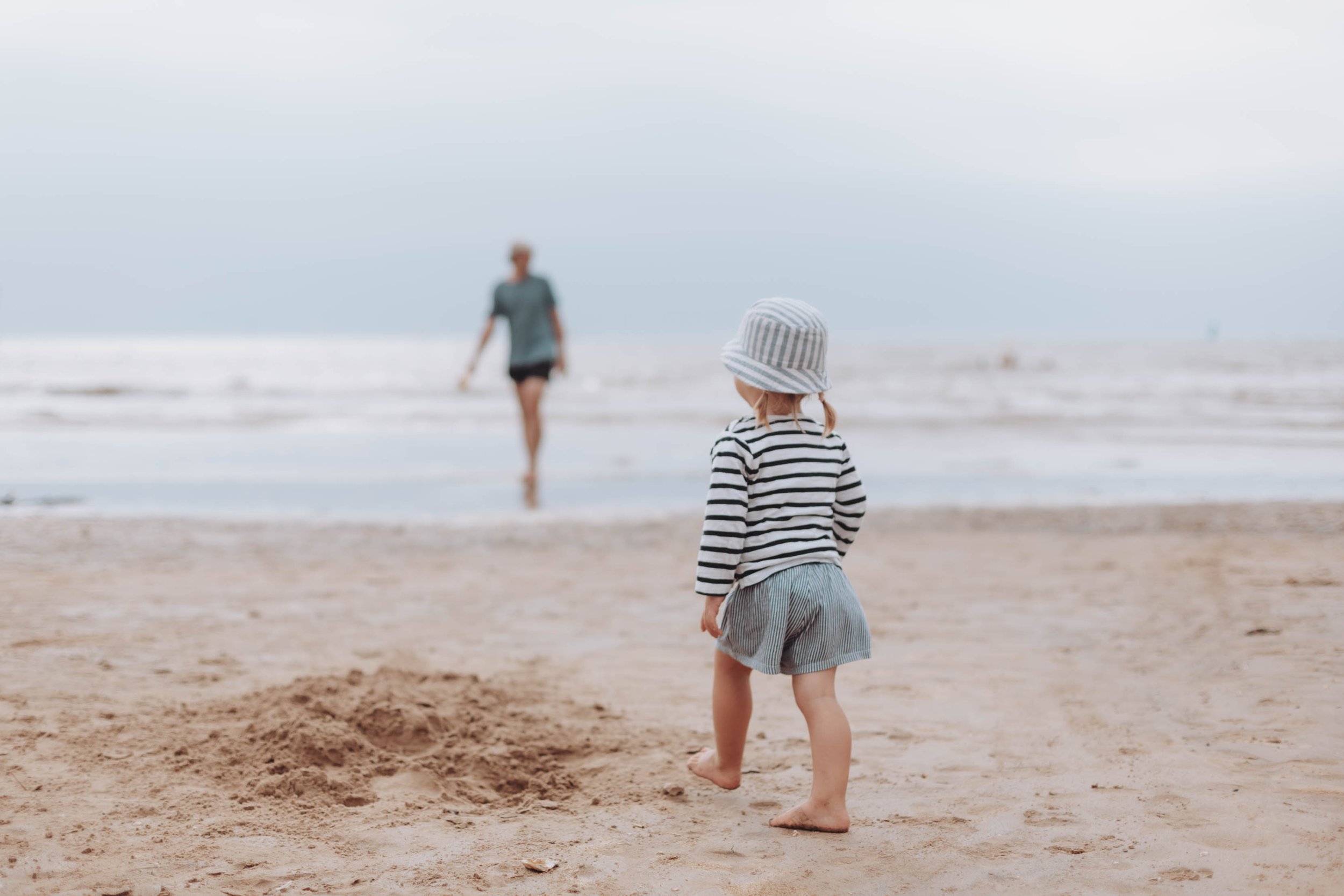 Un enfant en t-shirt rayé et short sur une plage, regardant vers l'eau où une personne marche au loin.