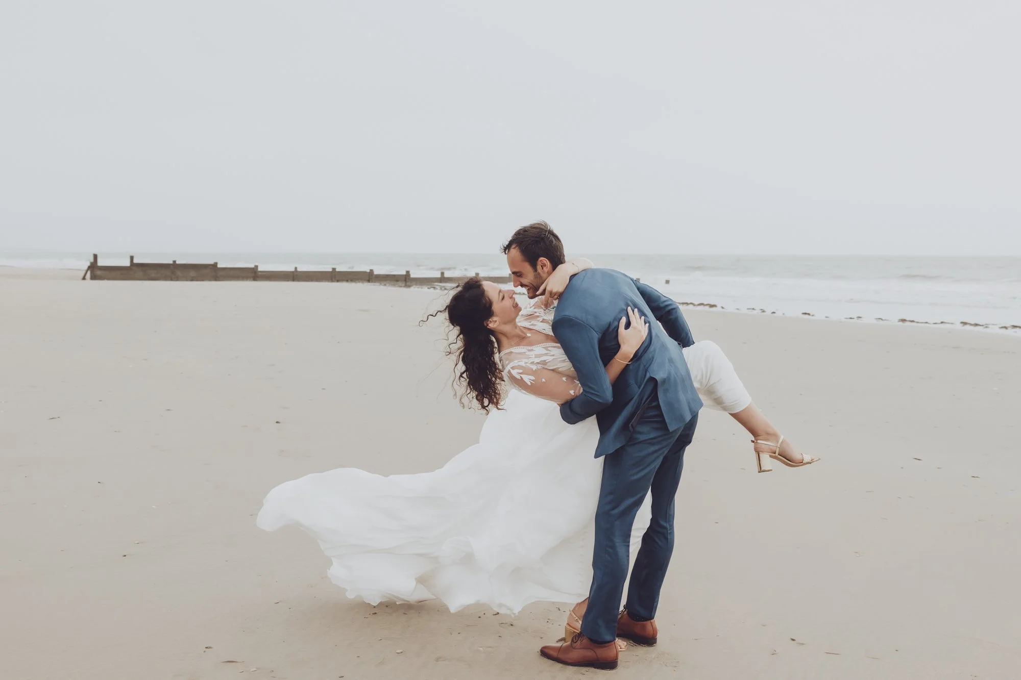 Un couple en tenue de mariage sur une plage, l'homme portant la femme dans ses bras, avec l'océan en arrière-plan.